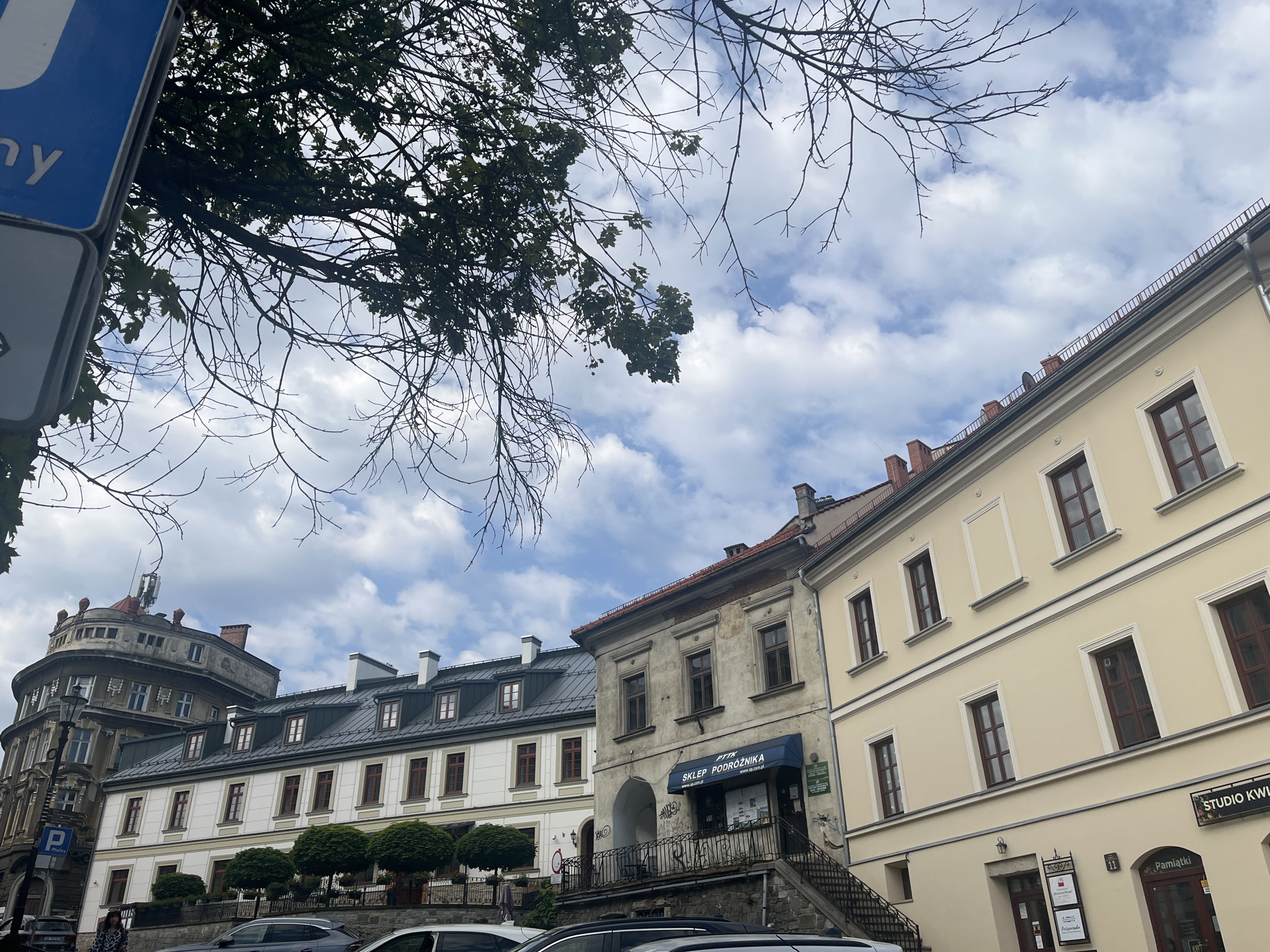 This media file captures an outdoor scene of a European street with historical architecture. The view is angled upwards, with trees and branches partially obscuring the sky, which is filled with white, puffy clouds against a blue background.
In the foreground, on the left, a blue and white sign is partially visible, with the letter "y" on the blue portion. Below this, a grey and white sign is also visible.
The main focus of the image is a row of buildings with distinct architectural styles. The building on the far left is a large, round, grey structure with multiple windows and an ornate top. Adjacent to it is a white building with a dark, sloped roof and several dormer windows. Further to the right is a lighter yellow building with a similarly dark, sloped roof. The closest building on the right is also yellow, with a dark tiled roof and visible chimneys. This building features a blue awning with white text that reads "SKLEP PODRÓŻNIKA". Below the awning, stairs lead up to an entrance. A sign to the right of this entrance reads "STUDIO KWI". Another sign further down the street indicates "Pamiątki".
There are a few people visible in the image, including one person in a patterned dress walking on the sidewalk in front of the white building. Two cars are parked on the street. The overall impression is of a sunny day in a historic town, possibly during daytime given the lighting. The presence of shops suggests a commercial area.