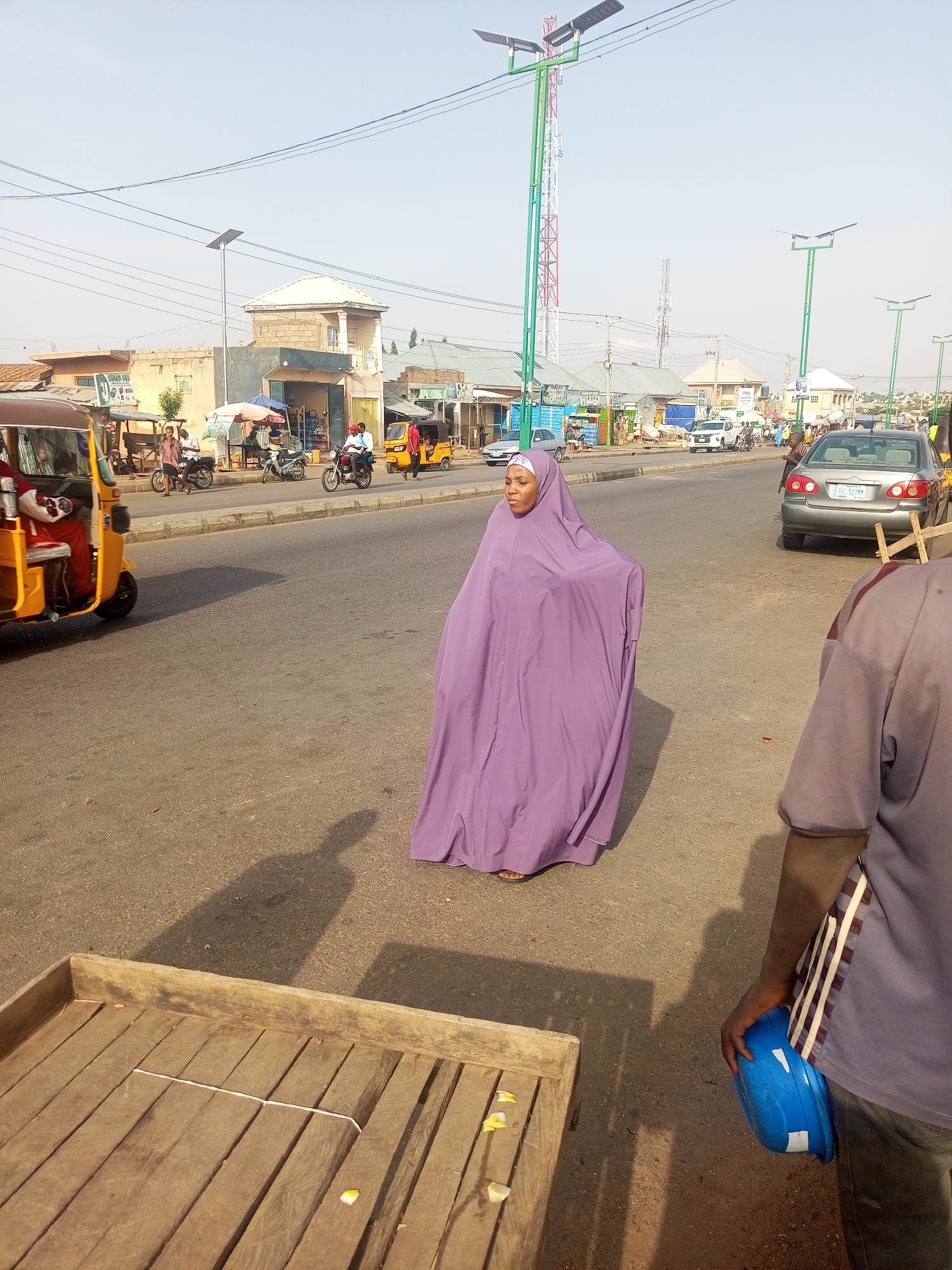 The image shows a street scene in what appears to be Tempure, Nigeria. A woman wearing a full-length, purple garment walks in the middle of the asphalt road. In the background, there are buildings, poles with solar panels, and traffic consisting of motorcycles, yellow auto rickshaws, and cars. A wooden cart is visible in the foreground on the left, and a man holding a blue object stands on the right side of the frame.