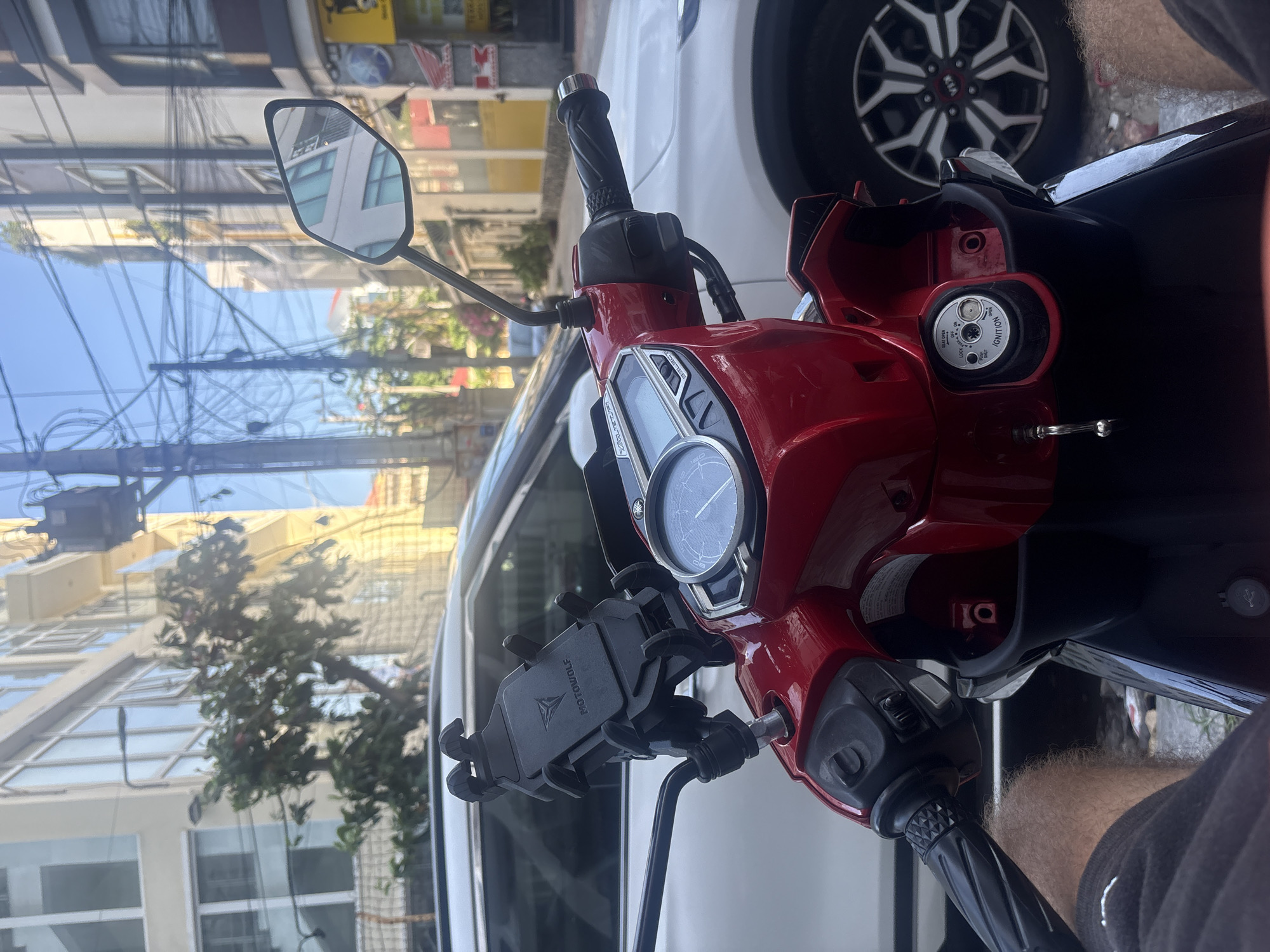 The image shows the front of a red motorcycle parked on a city street. The handlebars, instrument panel, and mirror are visible. A cell phone holder is mounted on the handlebars. A white car is parked next to the motorcycle. The reflection in the motorcycle mirror shows buildings, power lines, and a blue sky. The street appears to be in Bac Nha Trang Ward, Vietnam. The scene is captured from the rider's perspective. A person's arm wearing a black shirt is also visible.