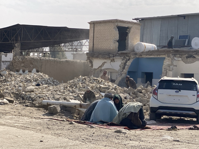 A group of men sits on a mat amidst rubble in front of a partially destroyed building. A white car is parked nearby. Debris is scattered across the foreground, with more destruction visible in the background, including a collapsed roof and damaged walls. Several men are observing from amidst the ruins. The scene appears to be in a region with war or natural disaster-related destruction.