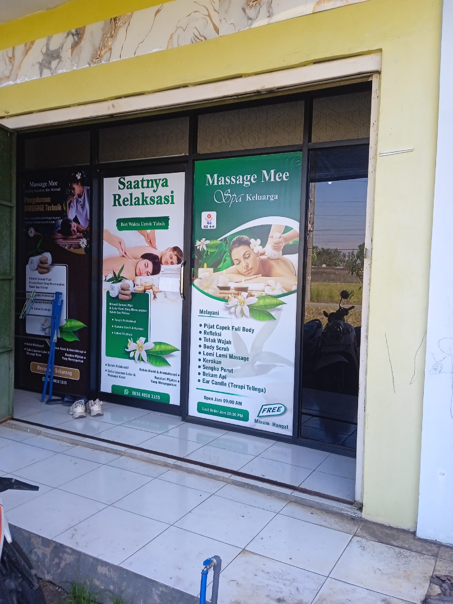 The image shows the facade of "Massage Mee Spa Keluarga," a massage business located in Cangkring, Indonesia. The business has a storefront with glass doors and large signs advertising its services, including massage, body scrubs, and ear candle therapy. One sign promotes "Saatnya Relaksasi" (Time for Relaxation). A motorcycle is parked outside the entrance, visible through the glass door. A pair of white shoes are visible on the floor. The facade is painted in light yellow and white, and the building appears to be in a commercial area.