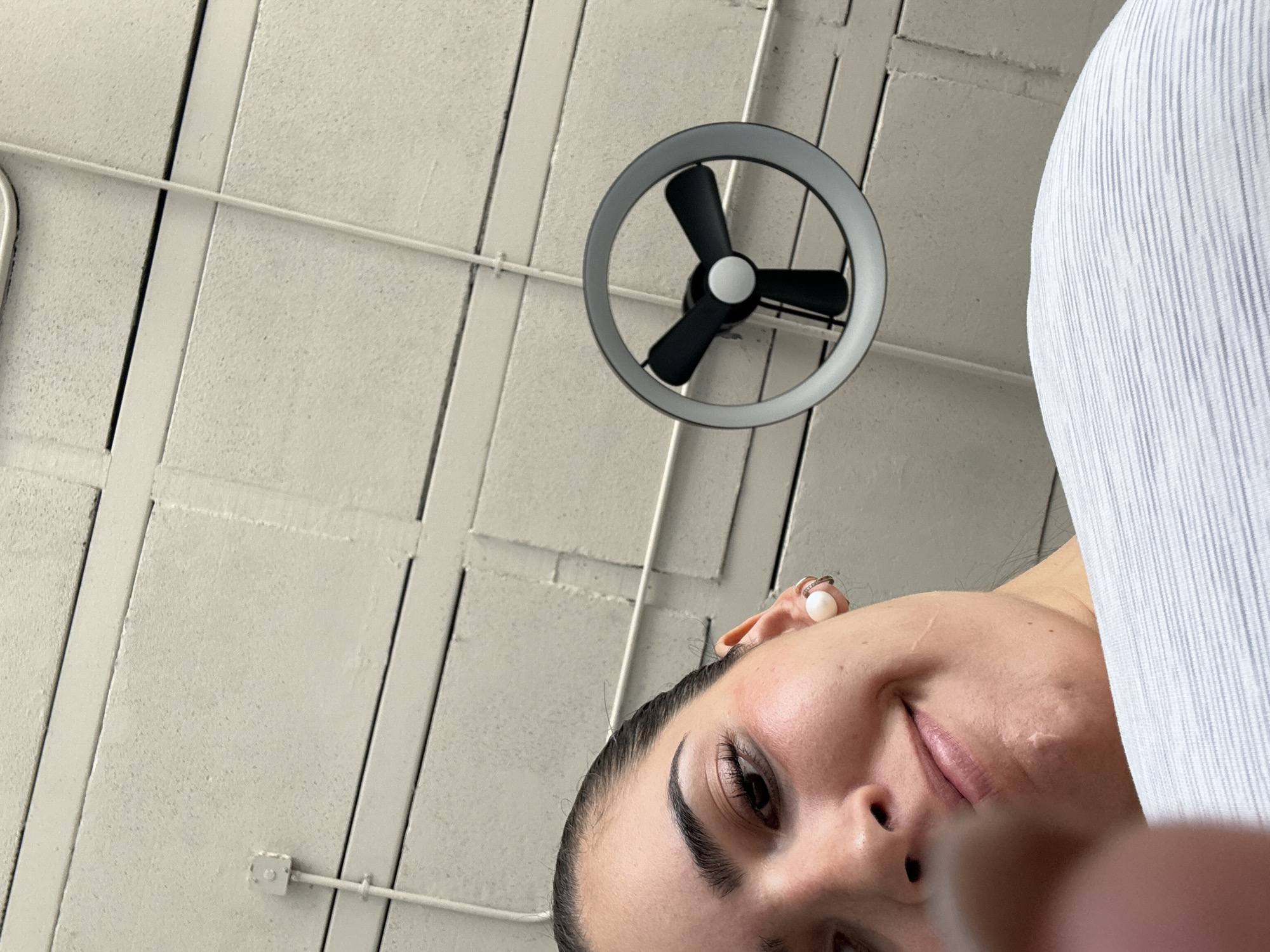 The image depicts a low-angle shot of a person's face, including their eye, nose, mouth, and right ear. The person has dark hair and is wearing what appears to be a white earbud in their right ear. The backdrop consists of a textured ceiling with recessed rectangular panels and a circular light fixture with three black blades, resembling a small fan. The person is wearing a white pinstripe shirt. The photo does not provide sufficient information to determine a specific location in Puebla, Mexico.