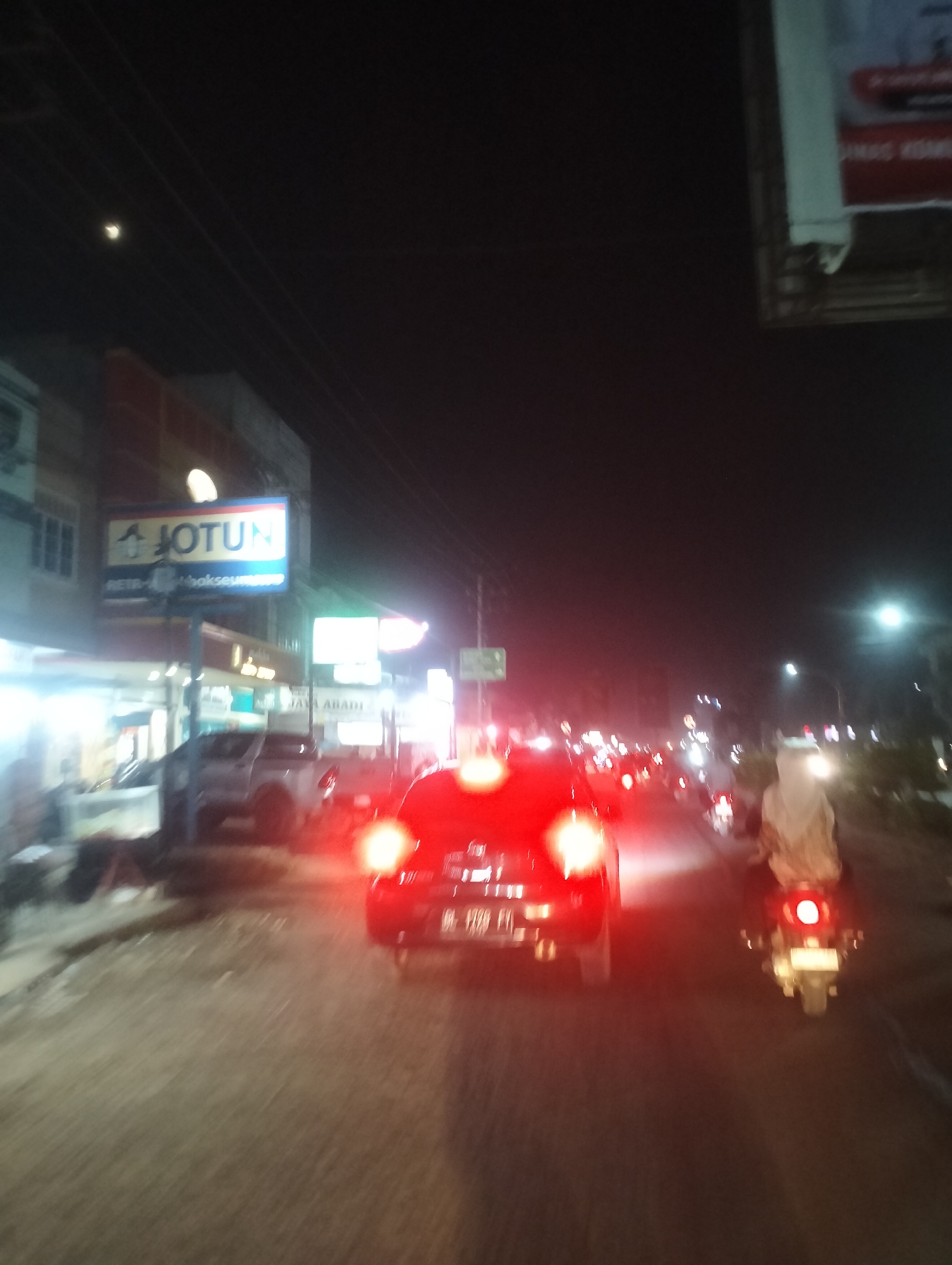 The image shows a street scene at night in Lhokseumawe, Indonesia. A bright red car is in the foreground, its taillights illuminated. To the right of the car, a person on a motorcycle is visible, also with their taillights on. The street is lined with buildings, one of which has a prominent "JOTUN" sign. There are other vehicles and lights visible in the background, suggesting moderate traffic. The overall lighting is dim, with artificial lights from street lamps and businesses illuminating the scene. The road surface appears to be asphalt.