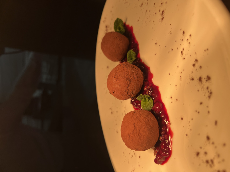 The image displays a beautifully plated dessert, likely chocolate truffles, presented on a white, slightly curved plate. Three spherical, dark brown items, coated generously with cocoa powder, are the central focus. These items are arranged diagonally across the plate, following the line of a vibrant red berry sauce or coulis.
The berry coulis is spread in a wavy, S-like pattern across the plate, starting from the top left and extending towards the bottom right. It appears to be made from berries like raspberries or cherries, as small fruit pieces and seeds are visible within the rich red liquid. Each of the three cocoa-dusted spheres is nestled alongside this berry coulis.
Fresh green mint leaves serve as a garnish, with small sprigs placed near each of the three dessert items, adding a touch of color and freshness. The white plate itself is clean, but a dusting of dark brown powder, similar to the coating on the spheres or cocoa, is scattered across the right side of the plate, enhancing the visual appeal.
The background on the left side of the image is very dark and out of focus, providing a stark contrast that makes the dessert stand out. There are faint, blurry reflections in the dark background, possibly from light sources or surrounding objects, but they are indistinguishable. The lighting in the image is warm and appears to come from the top or top-right, casting a soft glow on the dessert and highlighting its textures. This lighting suggests an intimate or refined setting, such as a restaurant or a special occasion. No text is visible in the image.