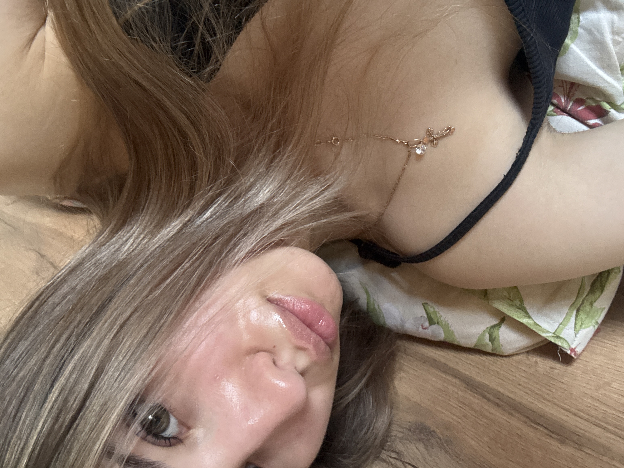 A close-up image shows a woman with light brown hair, part of her face visible. She is wearing a black top and a necklace with a cross pendant. She is lying on a bed with a patterned floral sheet, on top of a wooden floor. There is no evidence to determine the city or country.