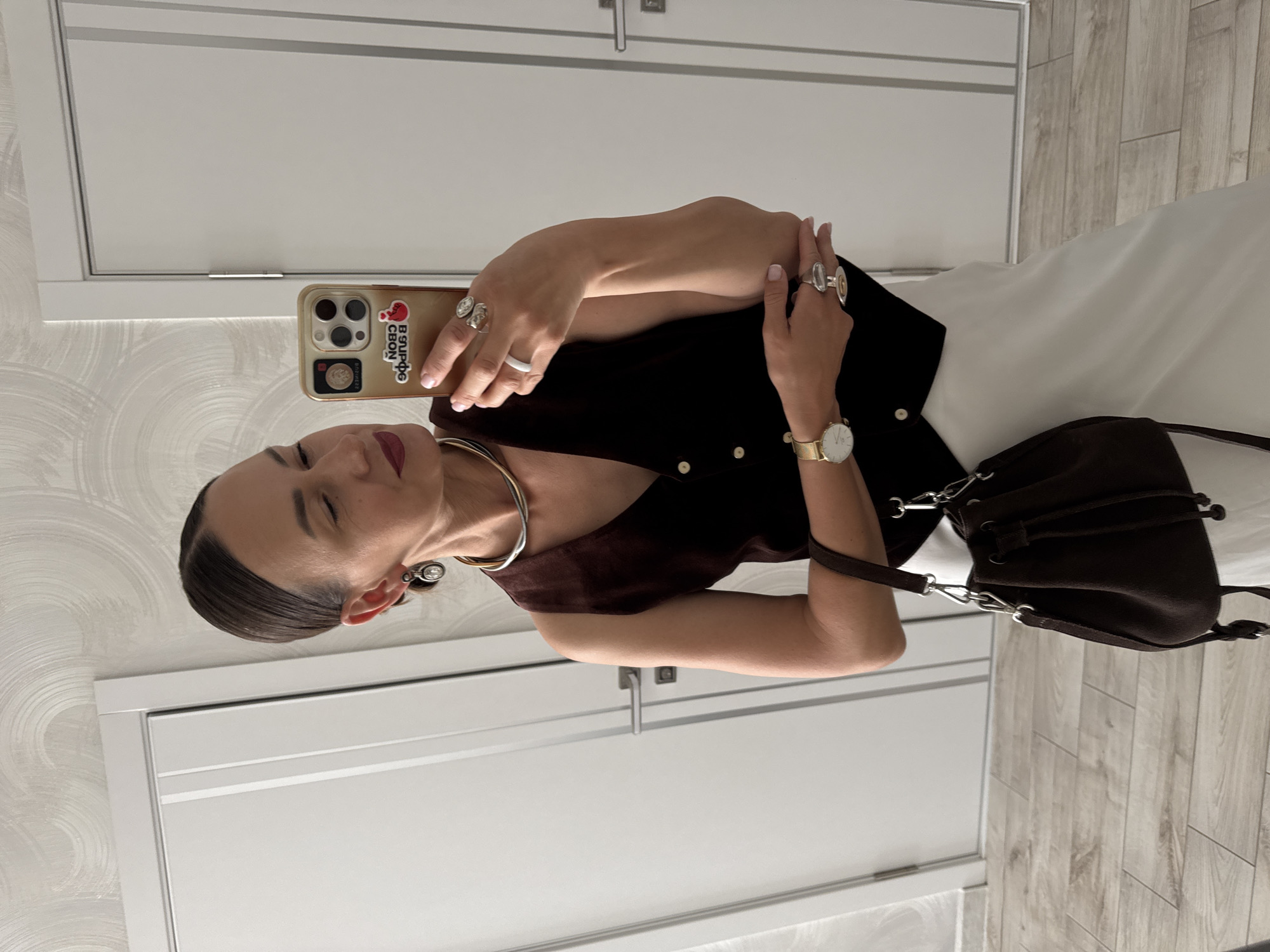 The image captures a woman taking a mirror selfie in what appears to be an indoor setting, possibly a home or hotel room in Sochi, Russia. The woman is dressed in a dark sleeveless top and a white skirt, accessorized with multiple rings on her fingers, a gold-toned necklace, a watch, and large earrings. She is holding a smartphone in her right hand, which has a case with several stickers, including one that appears to be in Cyrillic script. The background features white doors with horizontal detailing and a light-colored, patterned wallpaper. The floor is a light wood laminate. The lighting suggests it could be daytime, given the overall brightness of the scene, and there are no indications of weather. The woman's expression is neutral as she focuses on her phone. The overall impression is of a casual, everyday moment, with a focus on personal style and reflection.