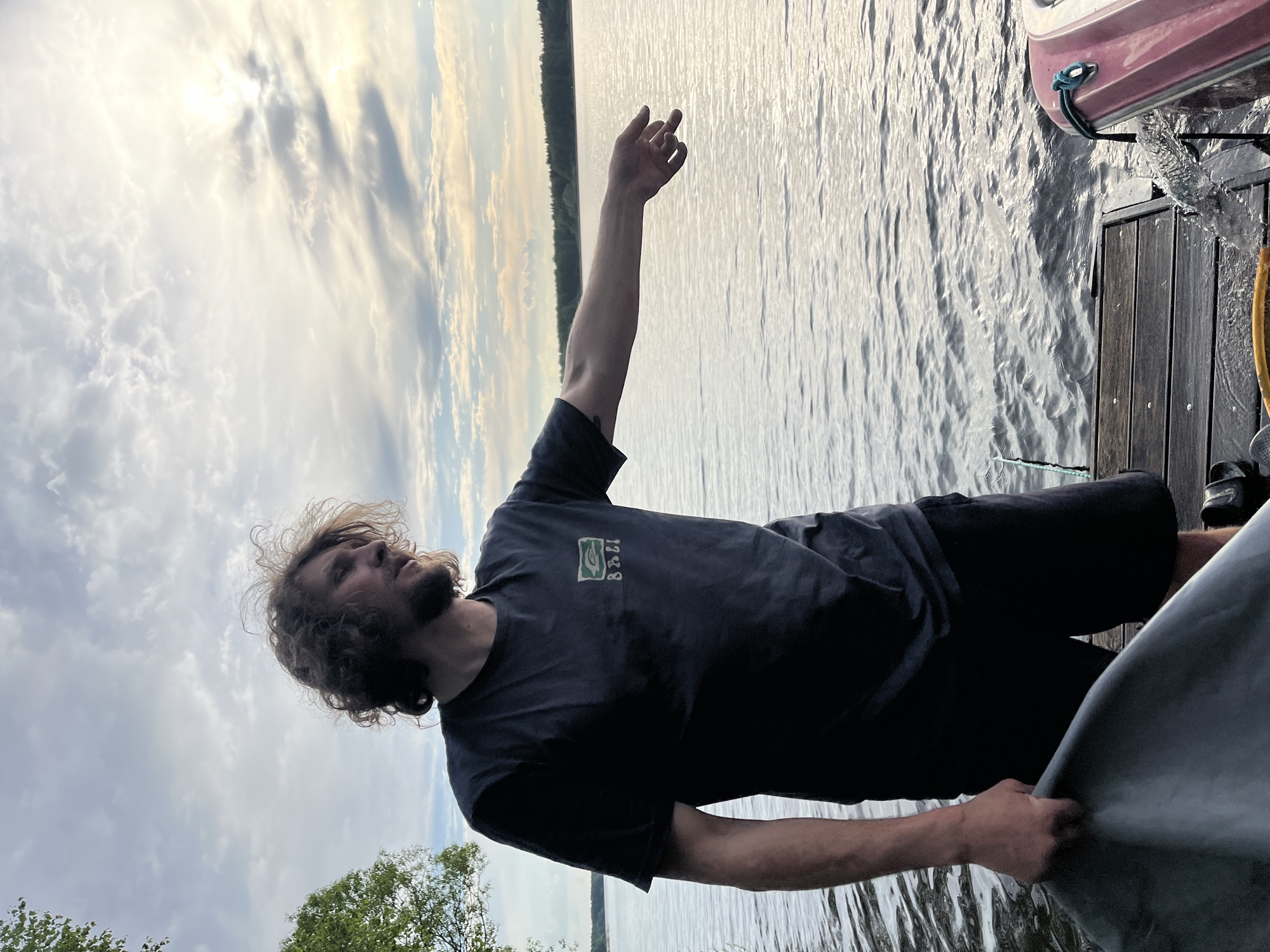A man with shoulder-length curly hair and a beard is standing on a wooden dock next to a body of water. He is wearing a dark t-shirt and shorts. His right arm is raised towards the sky. A boat is visible on the right side of the frame, partially on the dock. The sky is cloudy. There is green foliage visible on the left side of the frame, suggesting trees or shrubs. The image appears to be taken near Пятница, Russia.