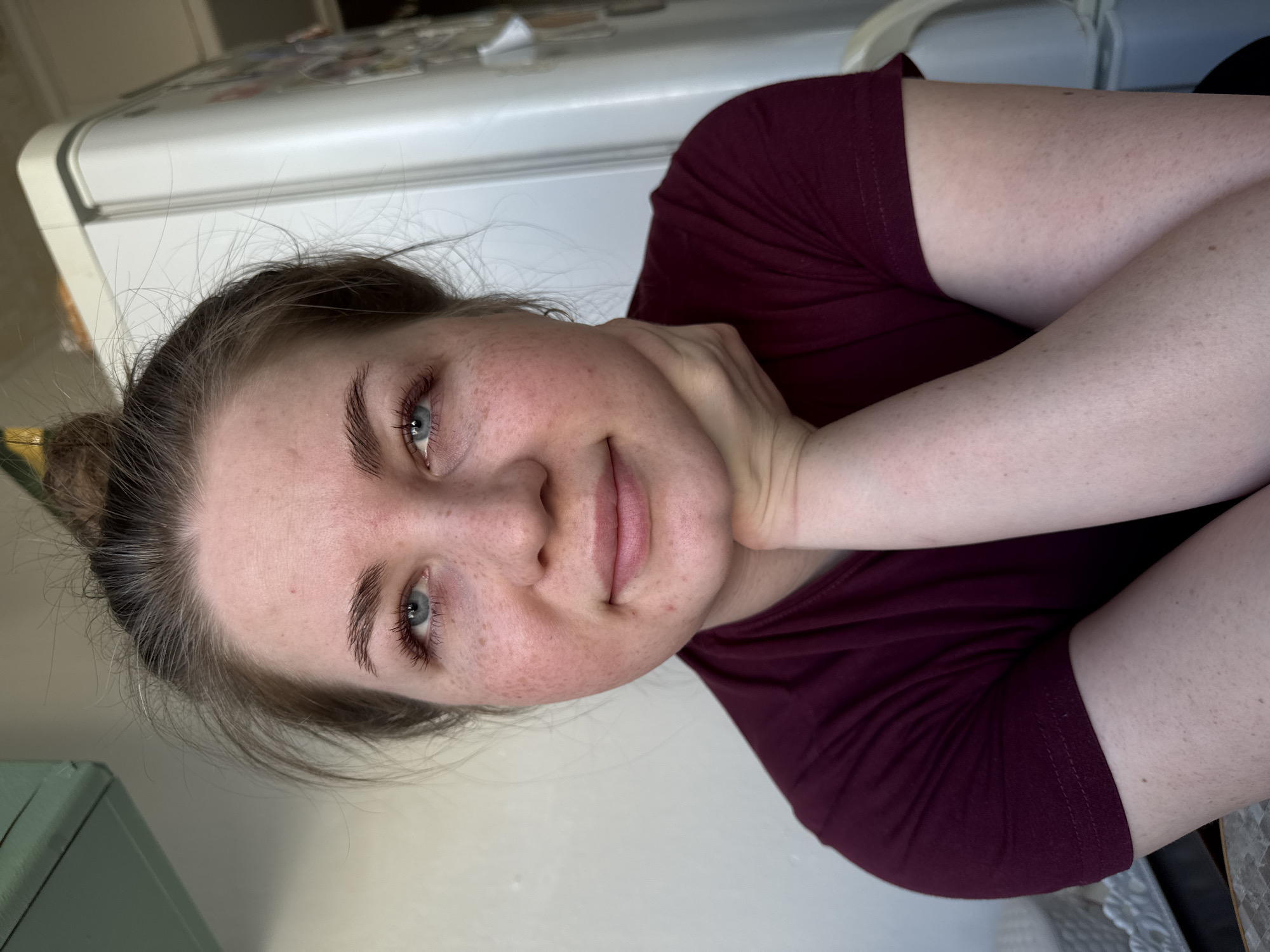A medium shot shows a fair-skinned woman with light brown hair pulled back in a messy bun. She has freckles on her face. Her eyes are blue and she is looking at the camera with a slight smile. She is wearing a maroon t-shirt and her right arm is bent with her hand supporting her cheek.
Behind her is a white refrigerator with magnets and papers stuck to it. A light-colored wall is to the right of the refrigerator. The edge of what appears to be a turquoise-colored cabinet or shelf is visible in the lower left corner. The location is not identifiable as Chaikovsky, Russia.