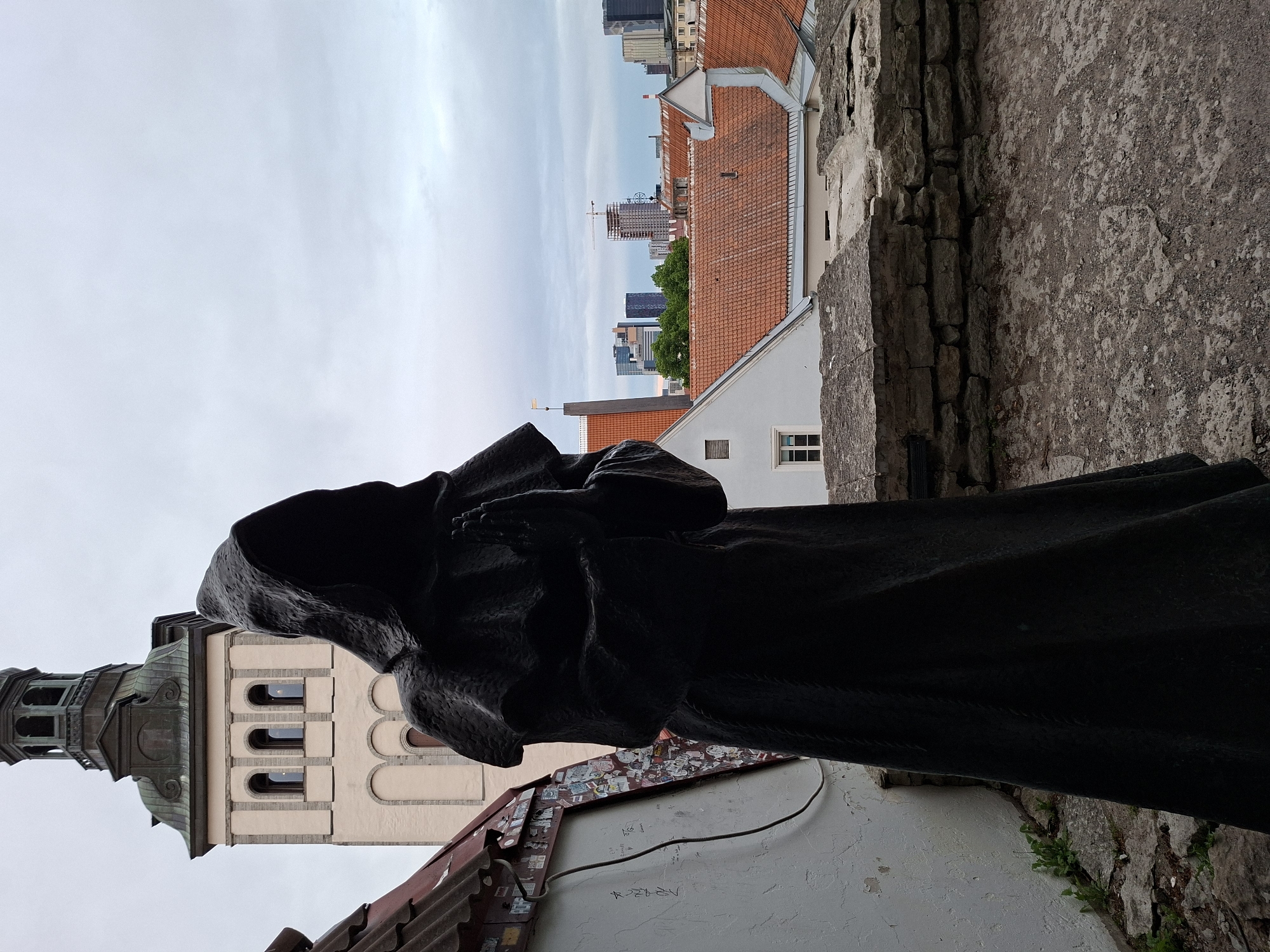 A statue of a cloaked figure, with hands clasped in prayer and a hood obscuring its face, stands in the foreground. Behind the statue, a light-colored tower with a dark, multi-tiered roof dominates the middle ground. The tower features arched windows and decorative elements. To the right of the tower, a panoramic view of Tallinn, Estonia, unfolds, showcasing a cityscape with red-tiled roofs, modern buildings, and a construction crane. The sky is overcast and pale gray. The foreground also includes a white wall adorned with numerous stickers and graffiti, a section of a red-tiled roof, and a low stone wall constructed from rough-hewn blocks. The ground is a textured, greyish surface.