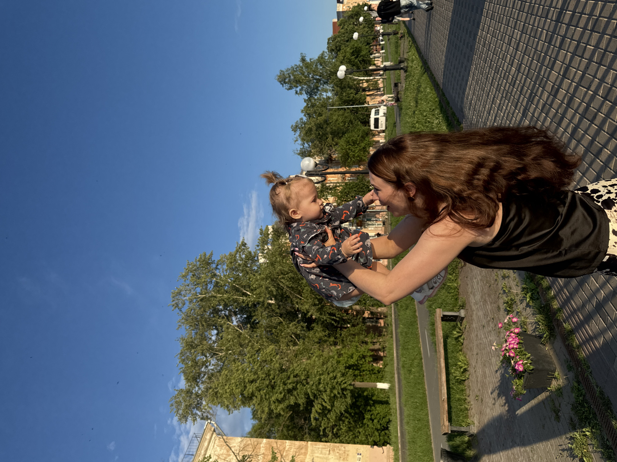 The image depicts a mother holding her baby outdoors on a sunny day. The mother, with long brown hair, is wearing a black top and patterned skirt. She is holding the baby up in the air, and they are looking at each other with smiles, suggesting a moment of joy and connection. The baby is wearing a patterned outfit and has their hair pulled up in a small ponytail.
The scene appears to be a park or a public walkway, with a paved path, green grass, and trees in the background. There are lampposts and a bench visible, along with some buildings in the distance. A white van can be seen parked on the side of the road. The sky is a clear blue, indicating fair weather. The sunlight casts shadows on the ground, suggesting it is daytime, likely late afternoon given the angle of the light.
The overall mood of the image is happy and relaxed, capturing a tender moment between a mother and her child in a pleasant outdoor environment. The location context of Votkinsk, Russia, provides a geographical anchor for the scene. There is no visible text in the image.