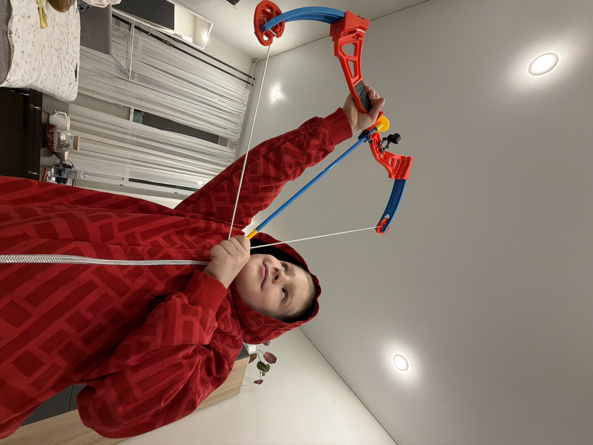 A child in a red hooded sweatshirt stands indoors, holding a toy bow and arrow. The location appears to be a domestic setting, potentially a bedroom. The room contains furniture such as a bed with bedding, a dresser, and a curtained window. The location is Засечное, Russia.