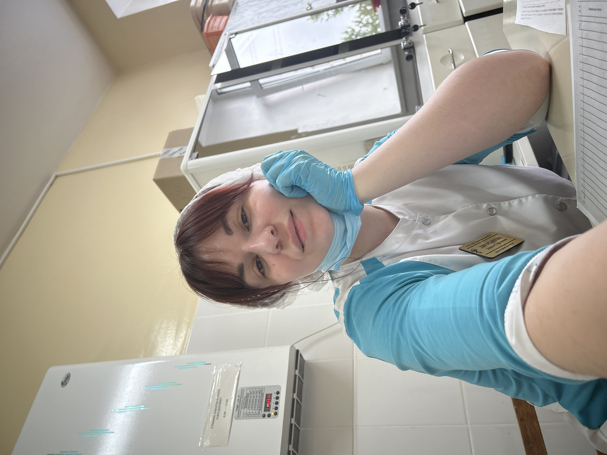 A woman in a medical uniform, consisting of a white scrub top with blue sleeves, a hairnet, and light blue surgical gloves, is taking a selfie in an indoor setting. She has reddish-brown hair, and a medical mask is pulled down below her chin. Her left hand rests on her cheek as she looks directly at the camera with a neutral expression.
The setting appears to be a medical or laboratory environment, with white tiled walls and a light-colored painted wall. Various pieces of equipment are visible: a large white appliance with a digital display showing "28" and some buttons is on the left. Above it, a sign with Russian text reads "ГРАФИК КВАРЦЕВАНИЯ" (Quartz Disinfection Schedule) and "ЕЖЕДНЕВНО" (Daily), with times like "9.00-15.00" and "10.0-18.0".
In the background, behind the woman, there's a piece of white and metallic furniture, possibly a laboratory bench or cabinet, and a window reflecting trees and a cloudy sky, indicating daytime. On the woman's scrub top, a nametag is visible with the text "ВРАЧ-ЛАБОРАНТ" (Doctor-Laboratory Assistant), "ДВОРНЕВА" (Dvorneva), and "Дарья Сергеевна" (Darya Sergeevna). To her right, a desk or counter with papers is partially visible. The image is rotated counter-clockwise.