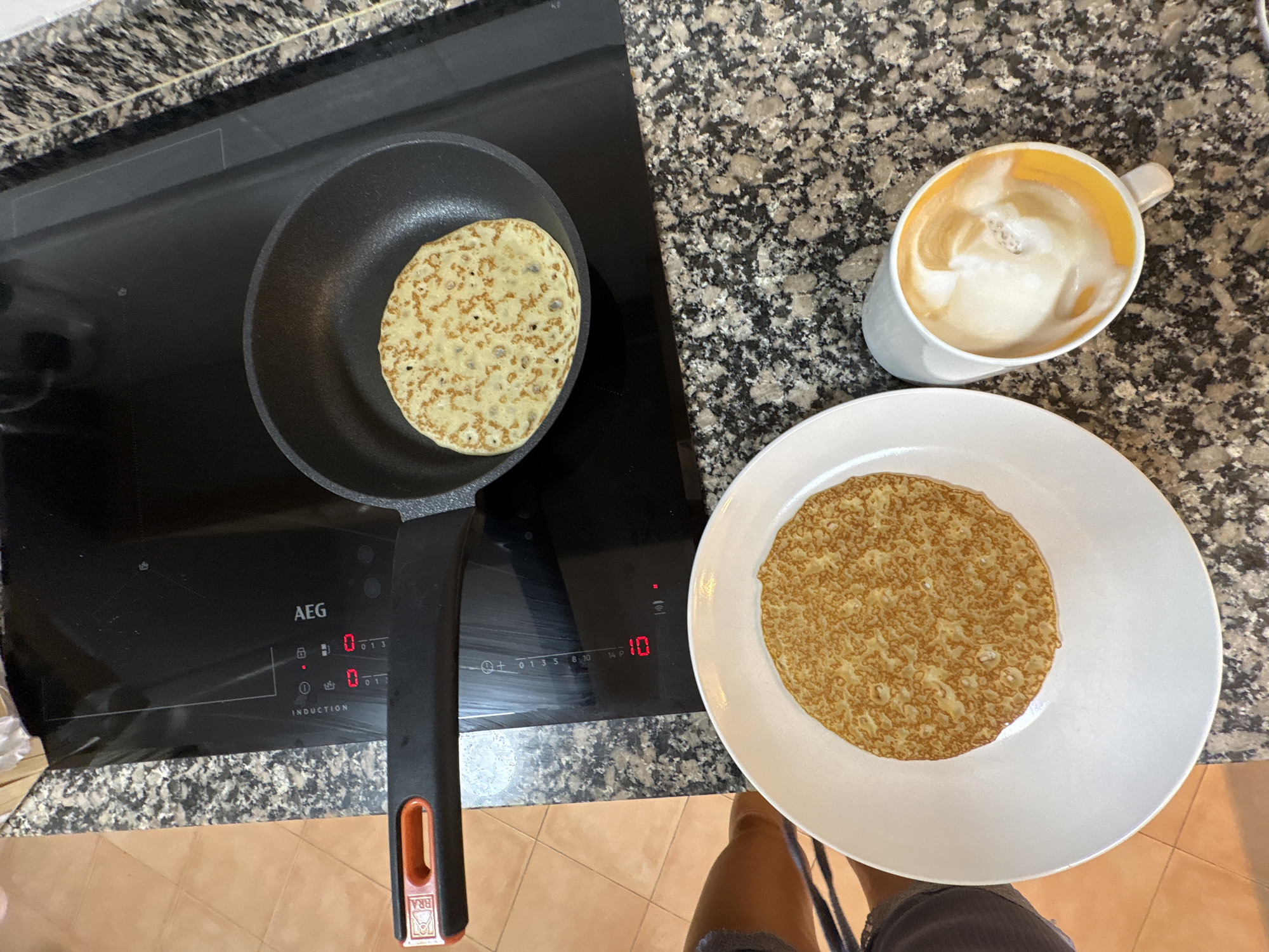 In an indoor setting, likely a kitchen, an induction cooktop is utilized for cooking. A black frying pan rests on the cooktop, containing a single, golden-brown pancake with a slightly textured surface. Beside the cooktop, a white plate holds another identical pancake. To the right of the plate, a white mug filled with a beverage topped with foamy milk and a swirl of amber liquid is positioned. The cooktop has a digital display showing "10" and "0". The overall scene suggests a breakfast or snack preparation. The background reveals a speckled granite countertop and a tiled floor, consistent with a domestic environment.