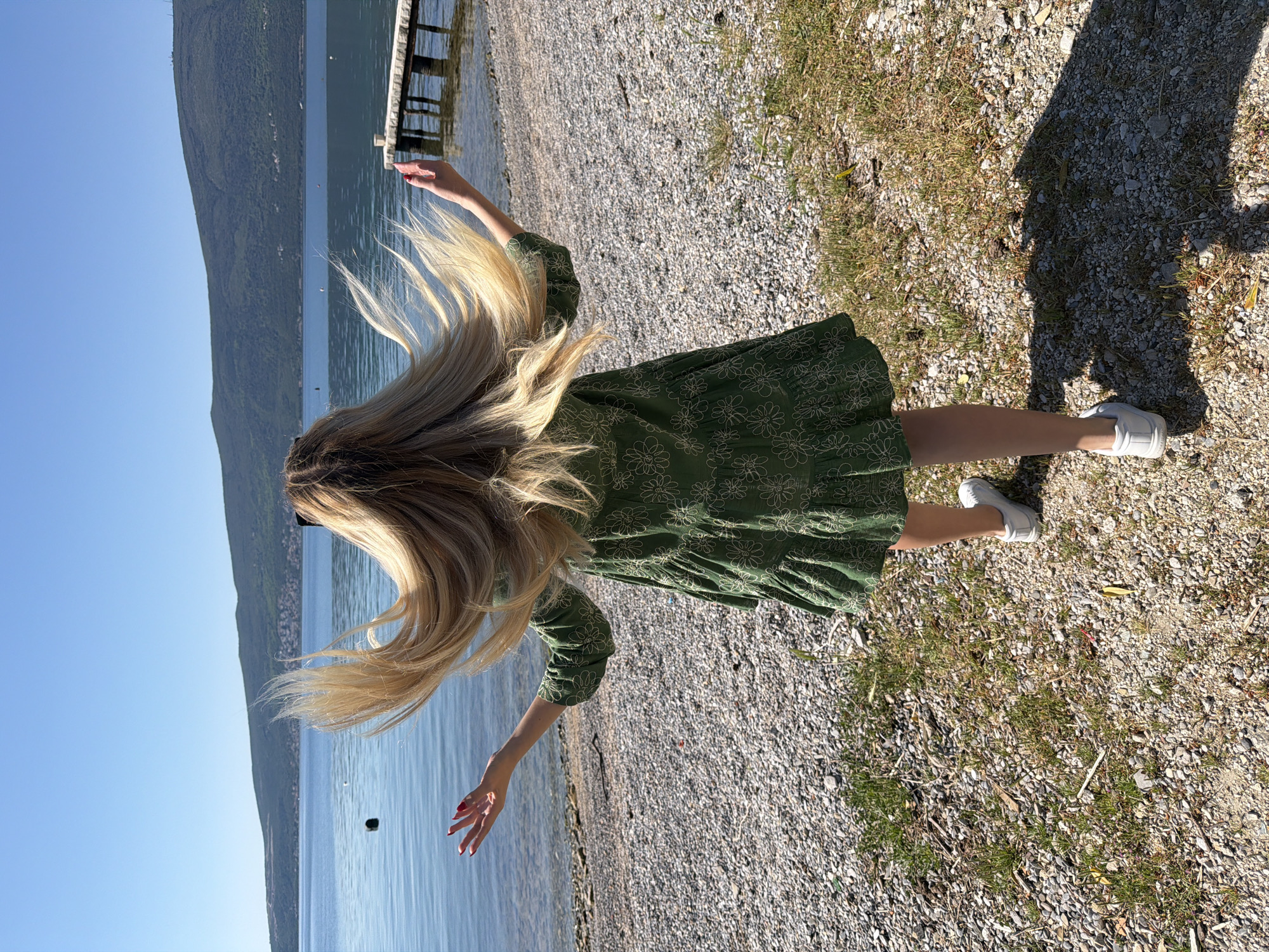 The image shows a person facing away from the camera, located on a pebble beach along the coastline. The person is wearing a green dress with floral embroidery and white sneakers, with long blonde hair. The arms are outstretched to the side. Behind the person is a body of water, likely the Bay of Kotor, with forested mountains in the background and a clear blue sky. To the right of the person is an area of ground with pebbles and patches of grass. A wooden pier can be partially seen in the background, extending into the water. Based on landmarks visible in the image, this location is in Bijela, Montenegro.