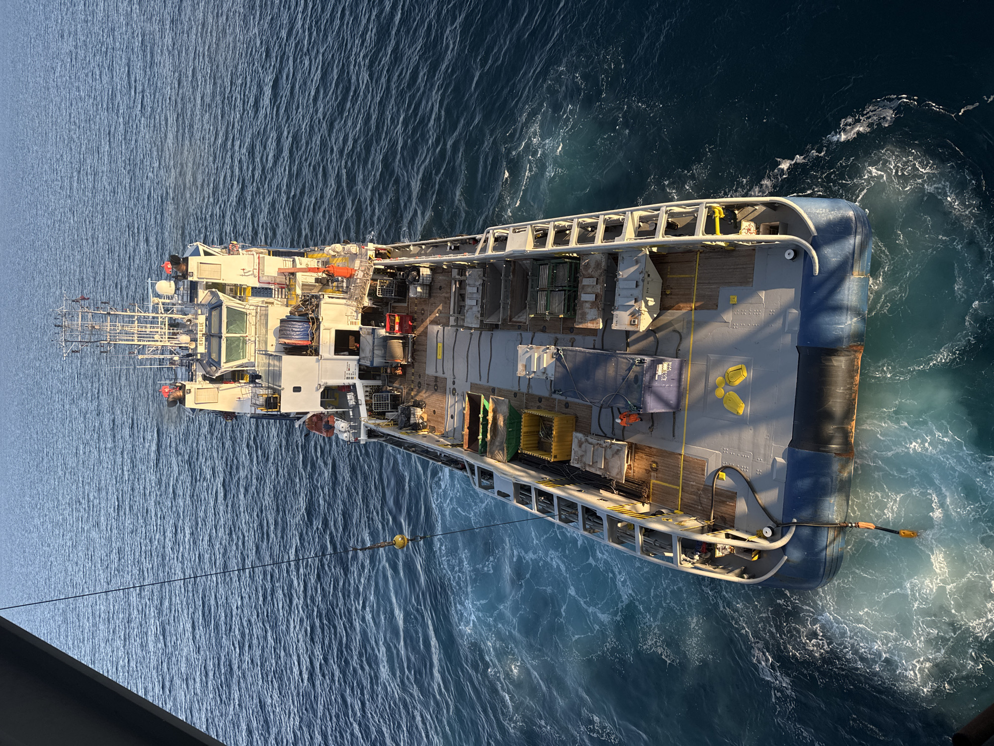 An aerial, high-angle, wide shot captures a commercial vessel in the water. The vessel is oriented diagonally from the top left to the bottom right of the frame. It has a white superstructure with multiple levels and a visible bridge at the front. The deck of the vessel is filled with various equipment, including yellow bins, green crates, and what appear to be pipes or poles. A crane arm extends from the left side of the image over the vessel, with a yellow, spherical object hanging from it. A person in an orange jumpsuit is visible on the deck near the stern of the vessel. The vessel is surrounded by dark blue water with white wakes and foam, indicating movement. The lighting suggests it is daytime, with sunlight casting shadows. The scene is outdoors on the open sea.