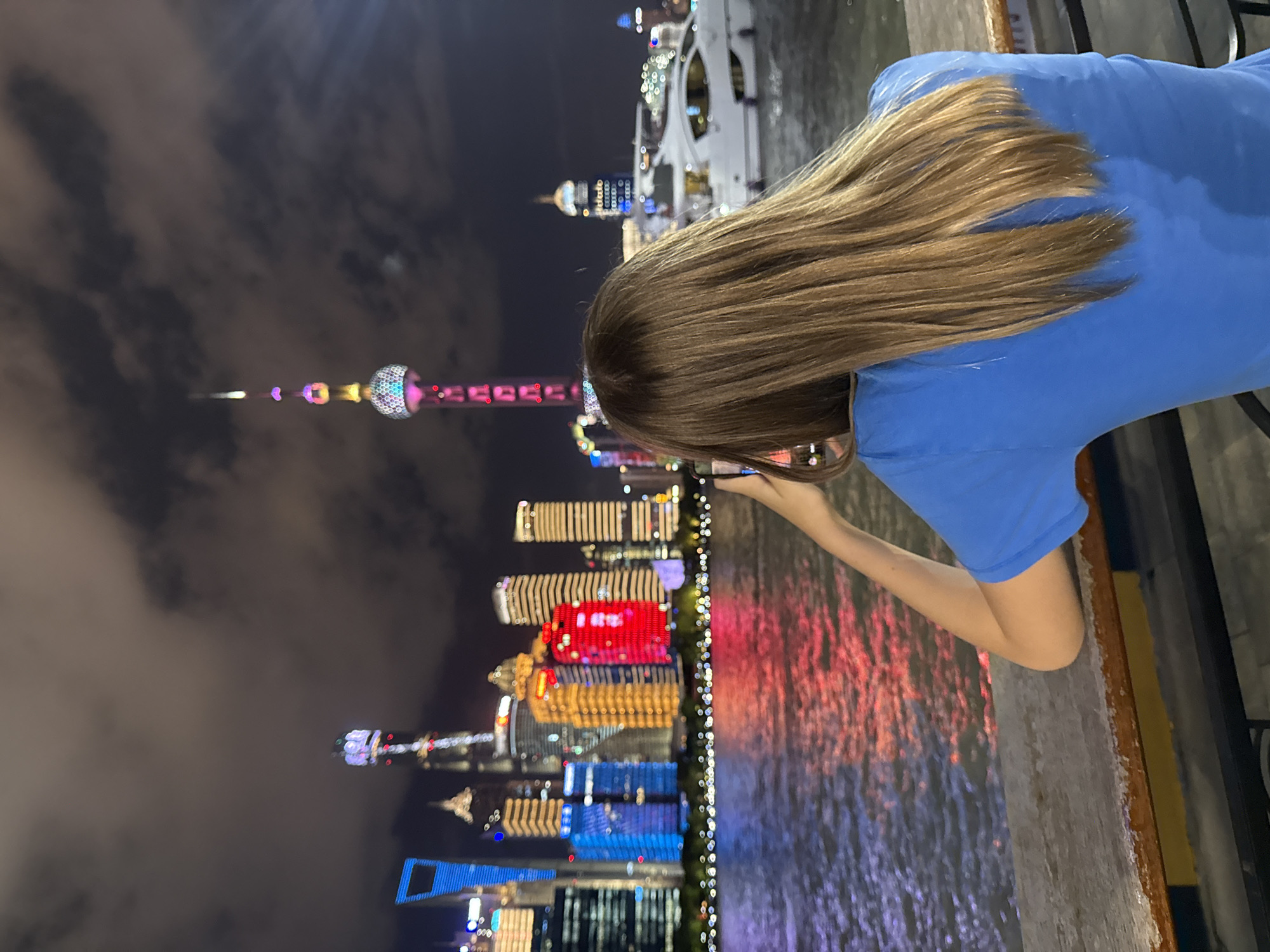 A person with long, light brown hair is viewed from behind, wearing a blue t-shirt, and holding a smartphone in their right hand, appearing to take a photo. They are standing on a walkway overlooking a body of water at night. Across the water, a cityscape is illuminated with colorful lights. Prominent structures include the Oriental Pearl TV Tower with its distinctive spheres and spire, and numerous skyscrapers with brightly lit facades in various colors, including red, yellow, and blue. The water reflects the lights of the city, creating a vibrant display. A white yacht is visible on the water to the right of the frame. The sky above is dark with clouds. This scene is likely the Bund waterfront in Hongkou District, Shanghai, China, known for its iconic skyline.