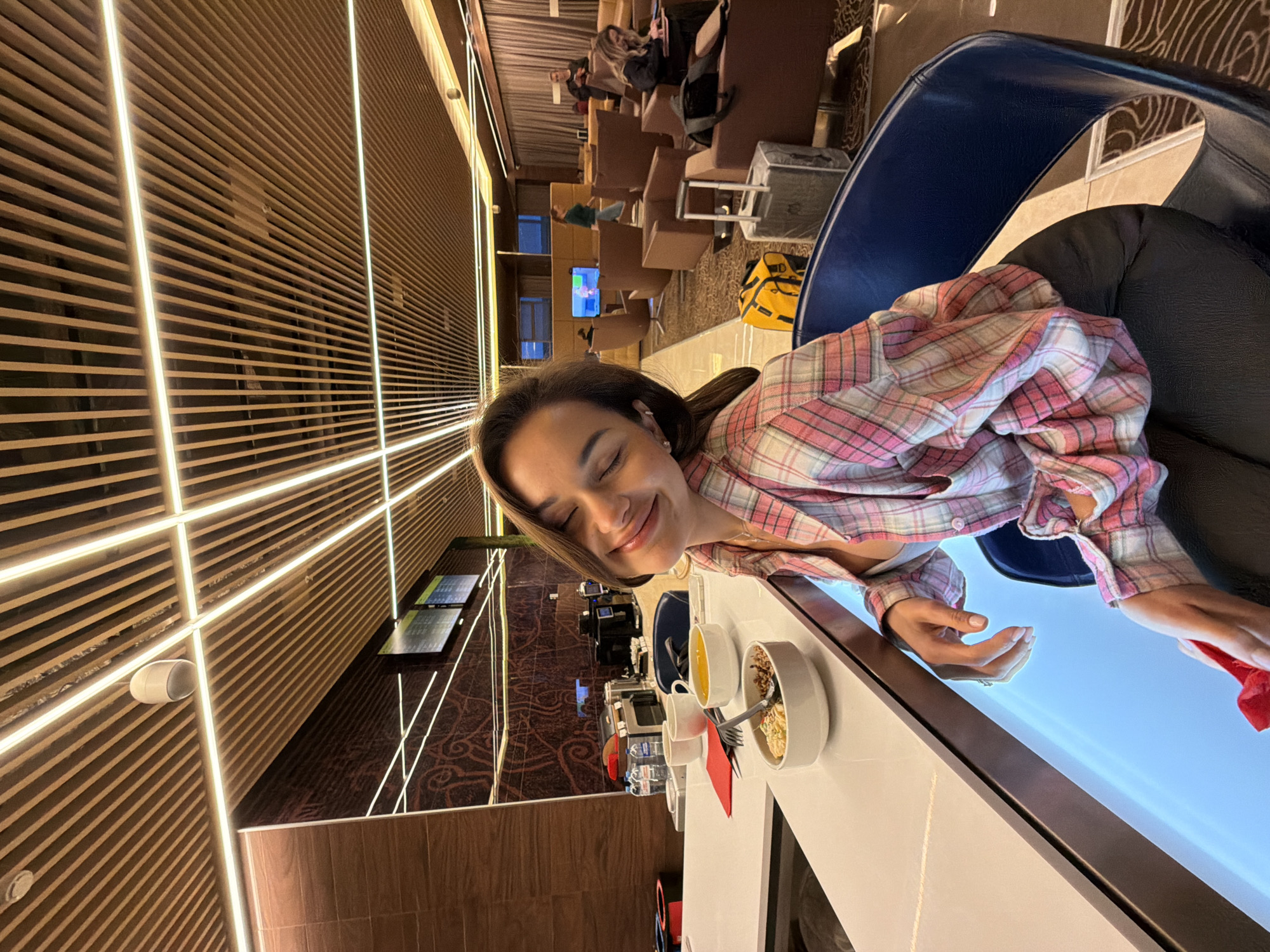 A woman is sitting at a table in what appears to be an airport lounge in the city of Unknown, Unknown. The woman is wearing a pink and white plaid shirt. On the table, there are several bowls and cups, possibly containing food and beverages. A red object is also visible on the table near her hand. The lounge has a modern design with wooden slats and illuminated lines on the walls. In the background, there are other people seated in lounge chairs, and television screens can be seen on a wall.