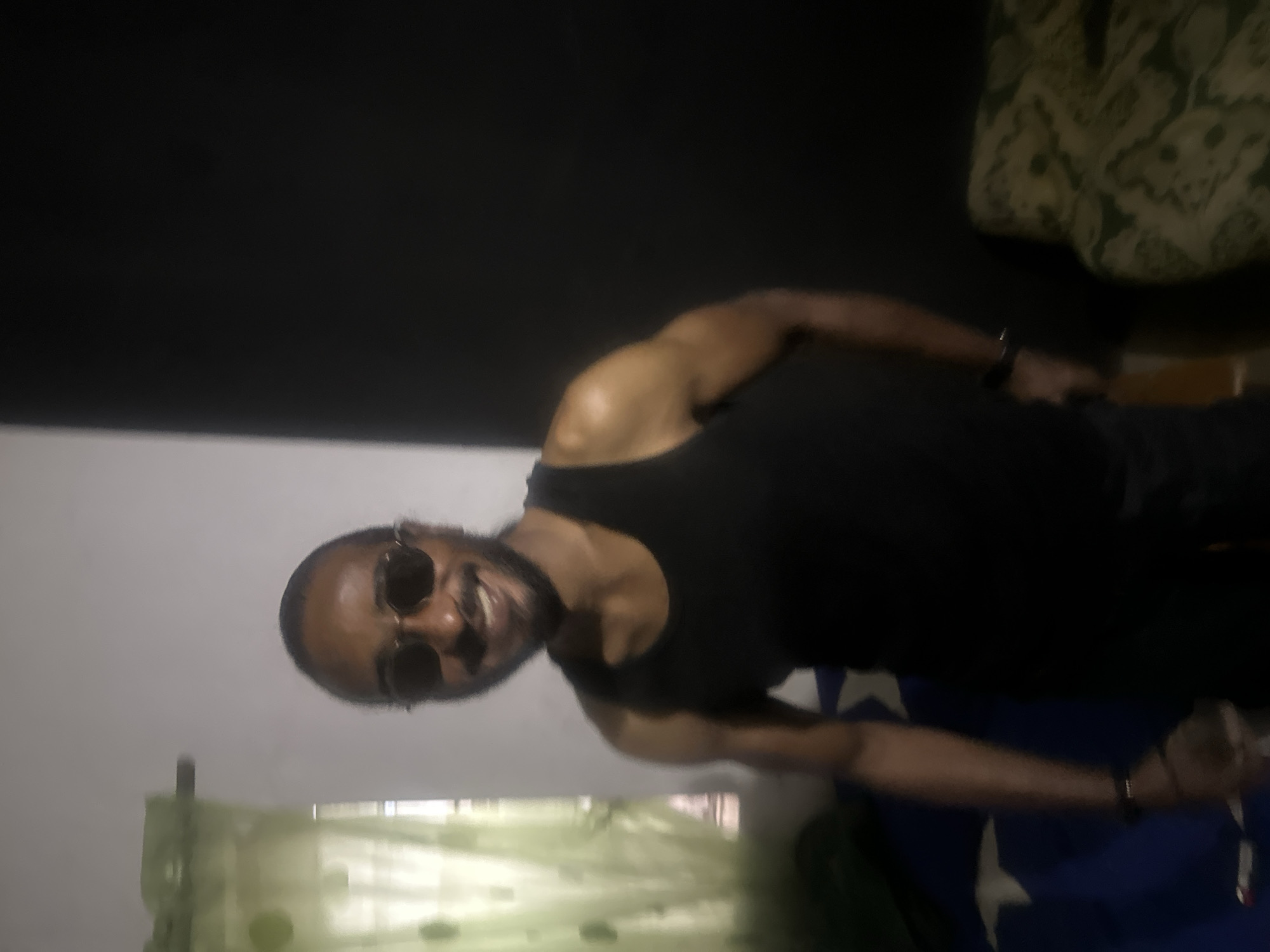 The image shows a man wearing a black tank top and sunglasses. He is smiling and looking directly at the camera. The background is blurred, but it appears to be an indoor setting with a window, curtains, and a bed with blue and white patterned sheets. Based on the prompt, the location is Jos, Nigeria, but this is not visible in the image itself.