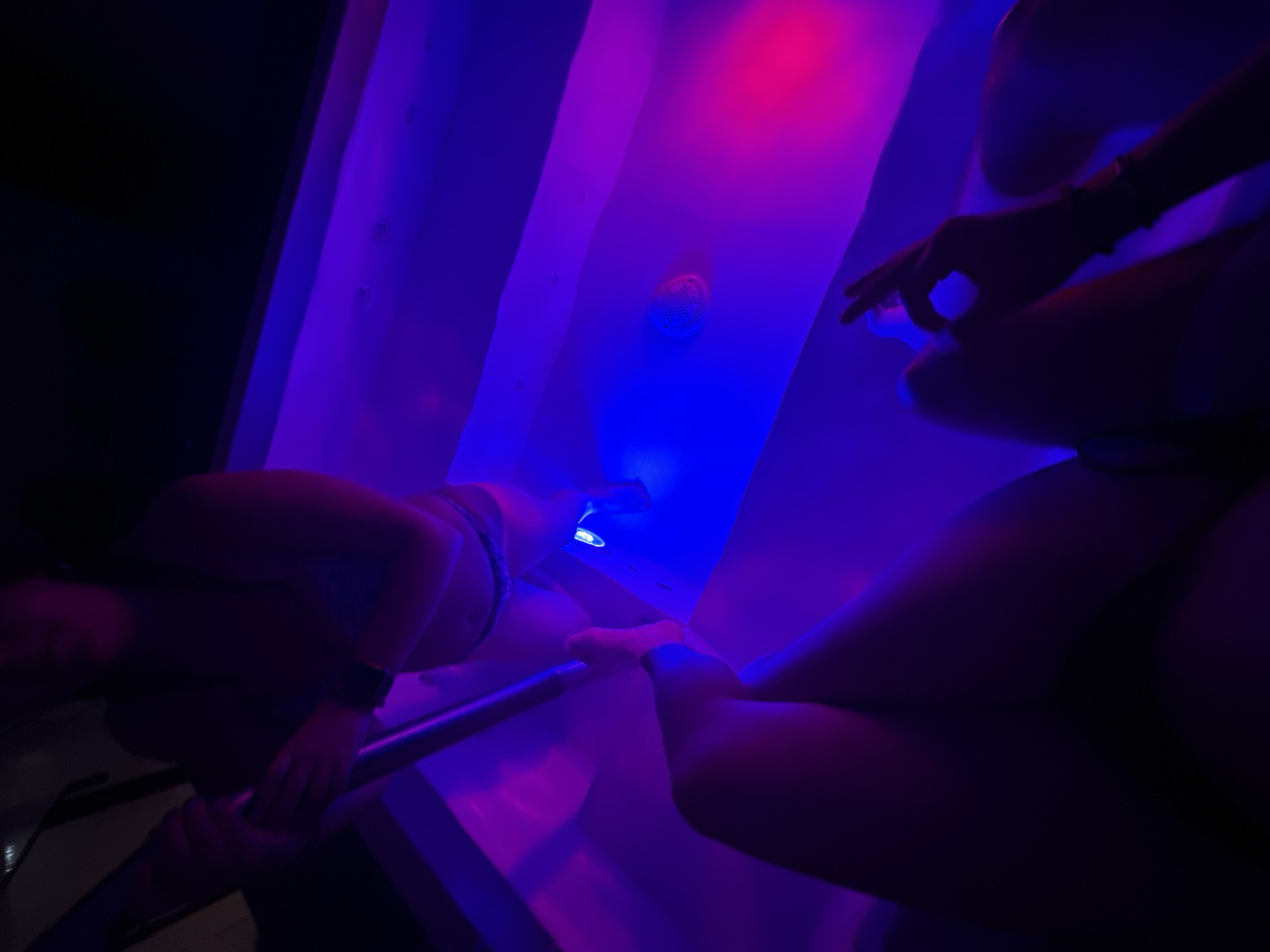 The image shows an indoor setting with two individuals, possibly in a sensory deprivation tank or a room with ambient lighting. The room is illuminated with a combination of blue and red lighting. One individual is seen partially submerged in what appears to be a tank or tub, holding onto a horizontal bar. Another individual is seen kneeling or sitting near the edge of the tank. The location is assumed to be Huixquilucan de Degollado, Mexico, but there are no distinct features that provide the exact location.