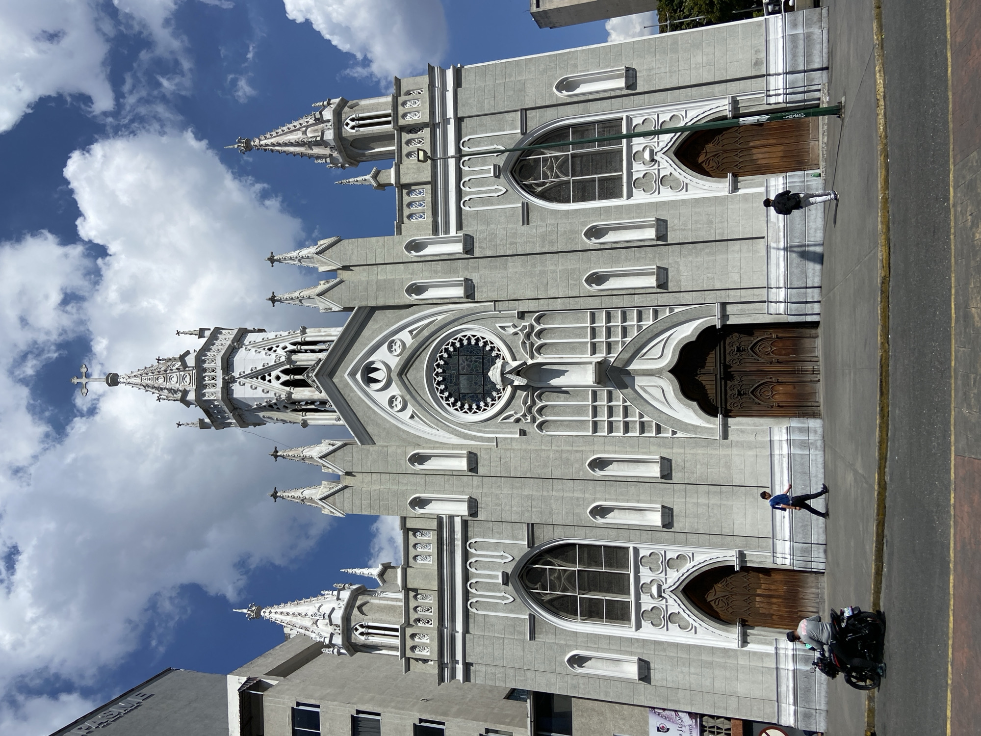 The image displays a full exterior view of the Santa Capilla Church in Caracas, Venezuela, a large grey Gothic Revival structure. The church features a central tall spire crowned with a cross, flanked by several smaller spires and pinnacles, all detailed with white accents. Its facade includes numerous pointed arches, a prominent circular rose window, and three large, carved wooden doors. The scene unfolds under a bright blue sky with scattered white cumulus clouds.
On the paved sidewalk in front of the church, three individuals are present. On the far left, a man wearing a grey long-sleeved shirt and a black cap is seated on a black motorcycle. To his right, a second man, dressed in a blue and white polo shirt and dark pants, walks from left to right. Further to the right, a third man, wearing a dark jacket and light-colored pants, walks away from the viewer. An urban street with a double yellow dividing line runs along the foreground. Adjacent grey buildings are visible on both the left and right sides of the church.