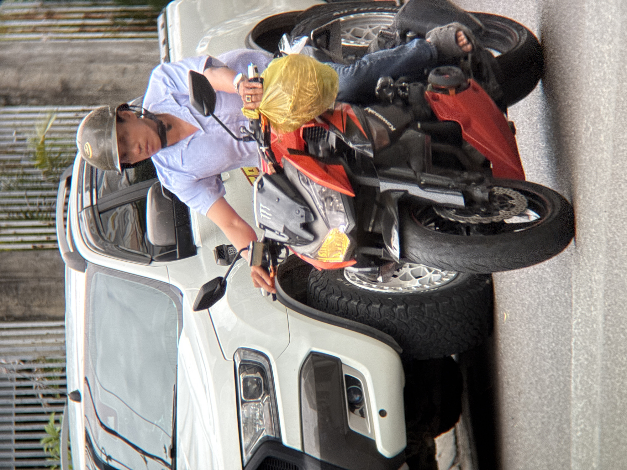 An individual wearing a light blue collared shirt, dark blue jeans, and a brown helmet rides a red and black motorcycle. The individual has a yellow plastic bag in their right hand. They are positioned on a paved road, with a white pickup truck to their left. The background includes a metal fence and greenery, suggesting an outdoor urban environment. The scene appears to be in Nha Trang, Vietnam, given the presence of a white pickup truck, common in the city, and the style of the motorcycle, consistent with vehicles found in Vietnam. The lighting suggests it is daytime, possibly overcast or late afternoon.