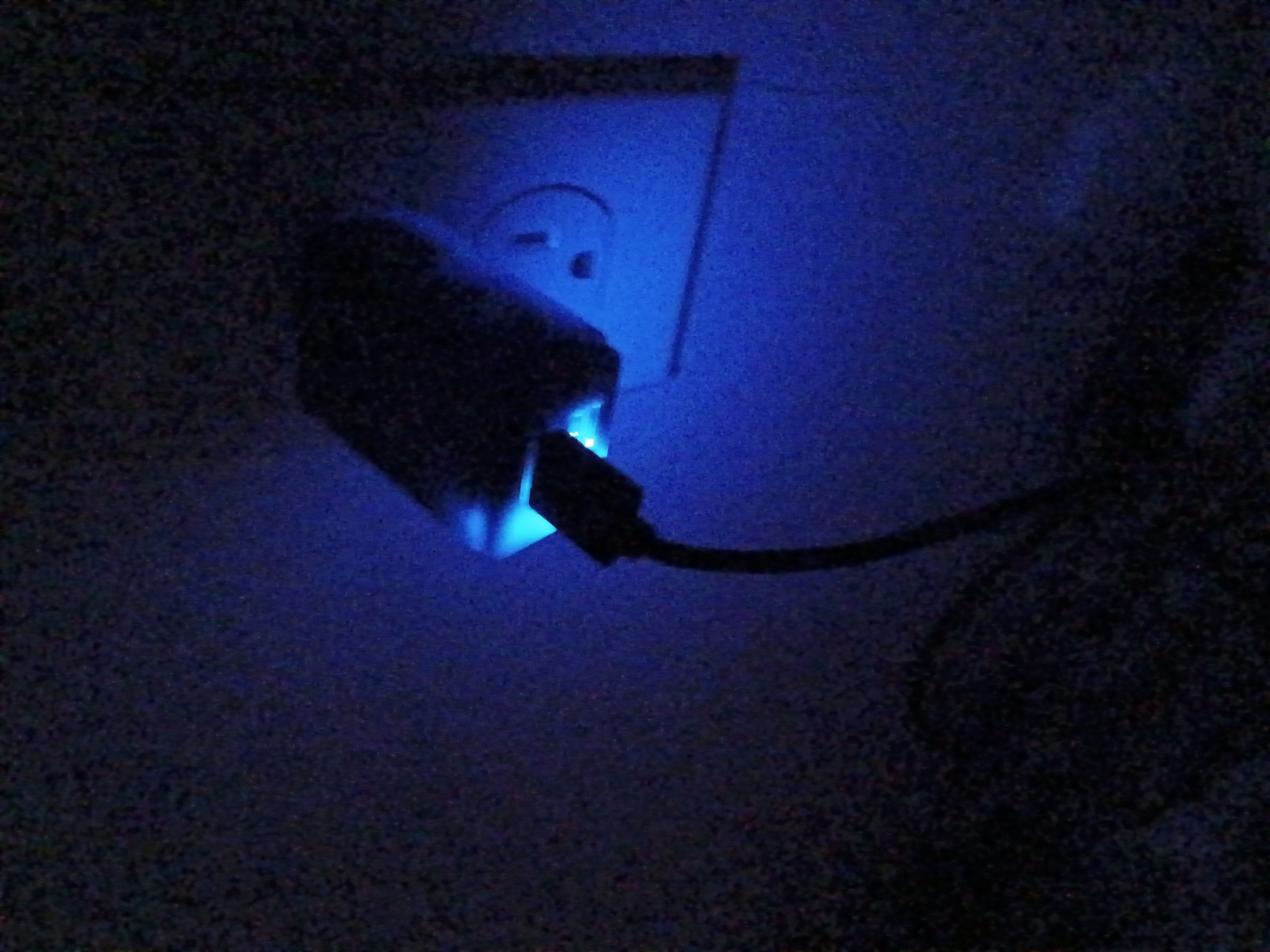 A low-light photograph shows a white electrical outlet with a USB charger plugged into it. A USB cable is attached to the charger. The light emitting from the charging device casts a blue hue on the scene. The location is not discernible from the image.