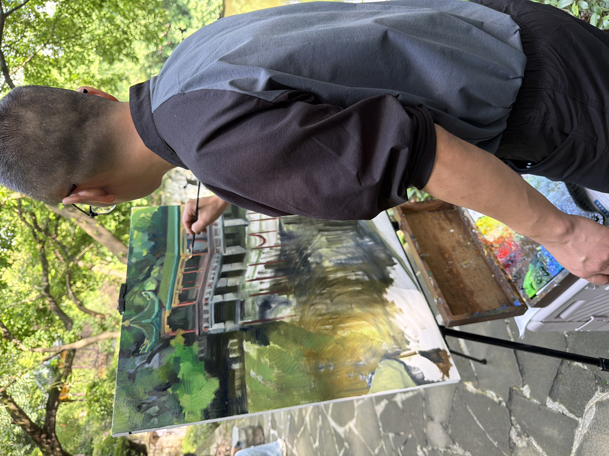 An artist paints a scene outdoors in Yuzhong District, China. The painting depicts a traditional Chinese pavilion with a green roof reflected in the water below, surrounded by lush greenery. The artist, a man wearing glasses and a dark and grey jacket over black pants, stands facing the canvas, holding a paintbrush and palette. An easel supports the painting, and a wooden paint box with various colors rests on a white portable container. The ground is paved with stone tiles, and the background is filled with the dappled light of trees.