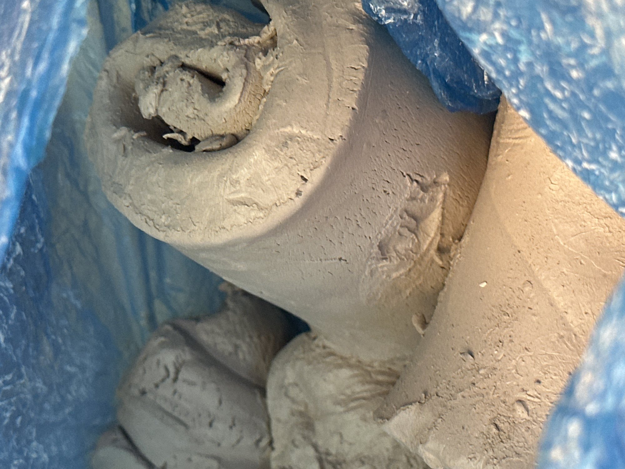 The image shows a close-up view of beige-colored clay material contained within a blue plastic bag. The clay appears to be formed into various shapes, some rolled or coiled, with rough, textured surfaces. The background is dominated by the blue plastic and the clay. The image is cropped tightly, obscuring any environmental context that might suggest a specific location within Nevinnomyssk, Russia. There are no people or discernible actions depicted.