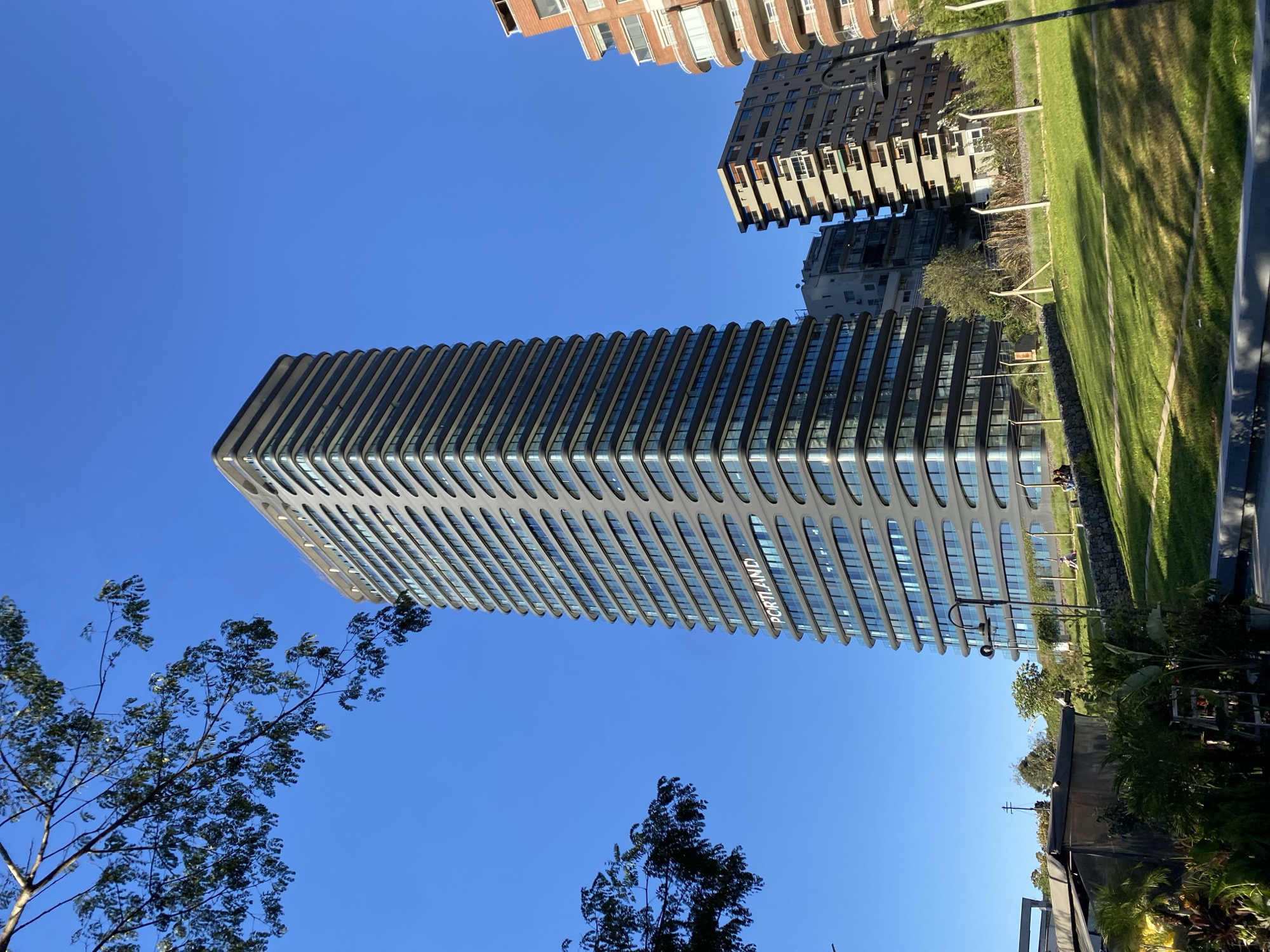The image shows the Alvear Tower in Buenos Aires, Argentina, with other buildings, trees, and a blue sky visible in the background. The tower has a modern design with repetitive horizontal elements. The word "PORTLAND" is visible on the building. A grassy area and some urban infrastructure are also present.