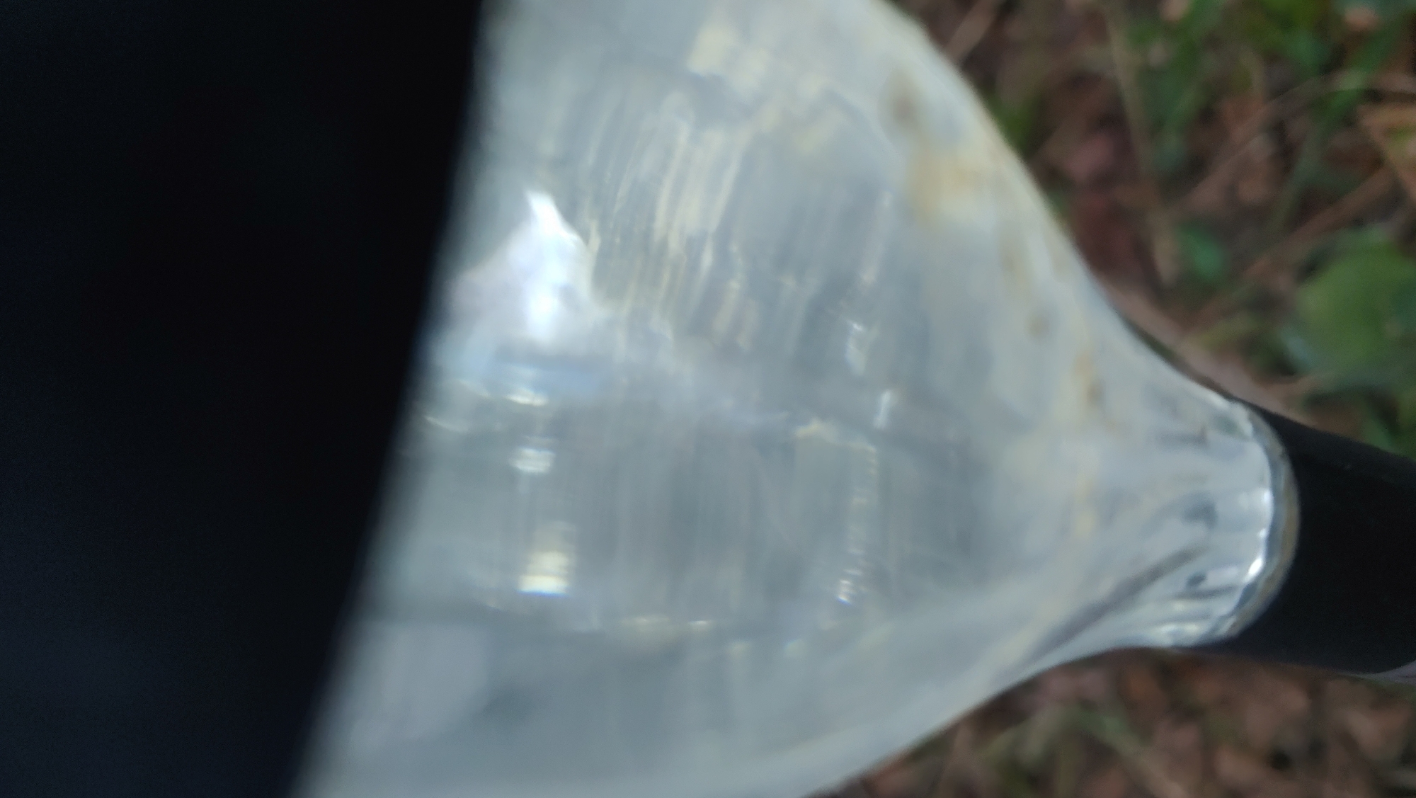 The media file is a close-up, abstract shot of a clear plastic bottle, possibly filled with liquid. The bottle is partially obscured by a black object on the left side of the frame, which could be part of a camera or another object. The bottle's surface shows internal ridges or textures, and light reflects off it, creating a shimmering, somewhat distorted effect. The background is out of focus, but appears to be an outdoor environment with elements of foliage and dry earth, suggesting a natural setting like a park or wooded area.
There are no people or identifiable text visible in the image. The lighting suggests it could be daytime, but the specific time is not discernible. The overall impression is one of natural light interacting with a transparent object in an outdoor environment. The image is devoid of any specific activity or event, focusing solely on the visual qualities of the bottle and its interaction with light and its surroundings. The location context of Mukachevo, Ukraine, does not provide specific visual cues within the frame itself, but adds a geographical layer to the description.