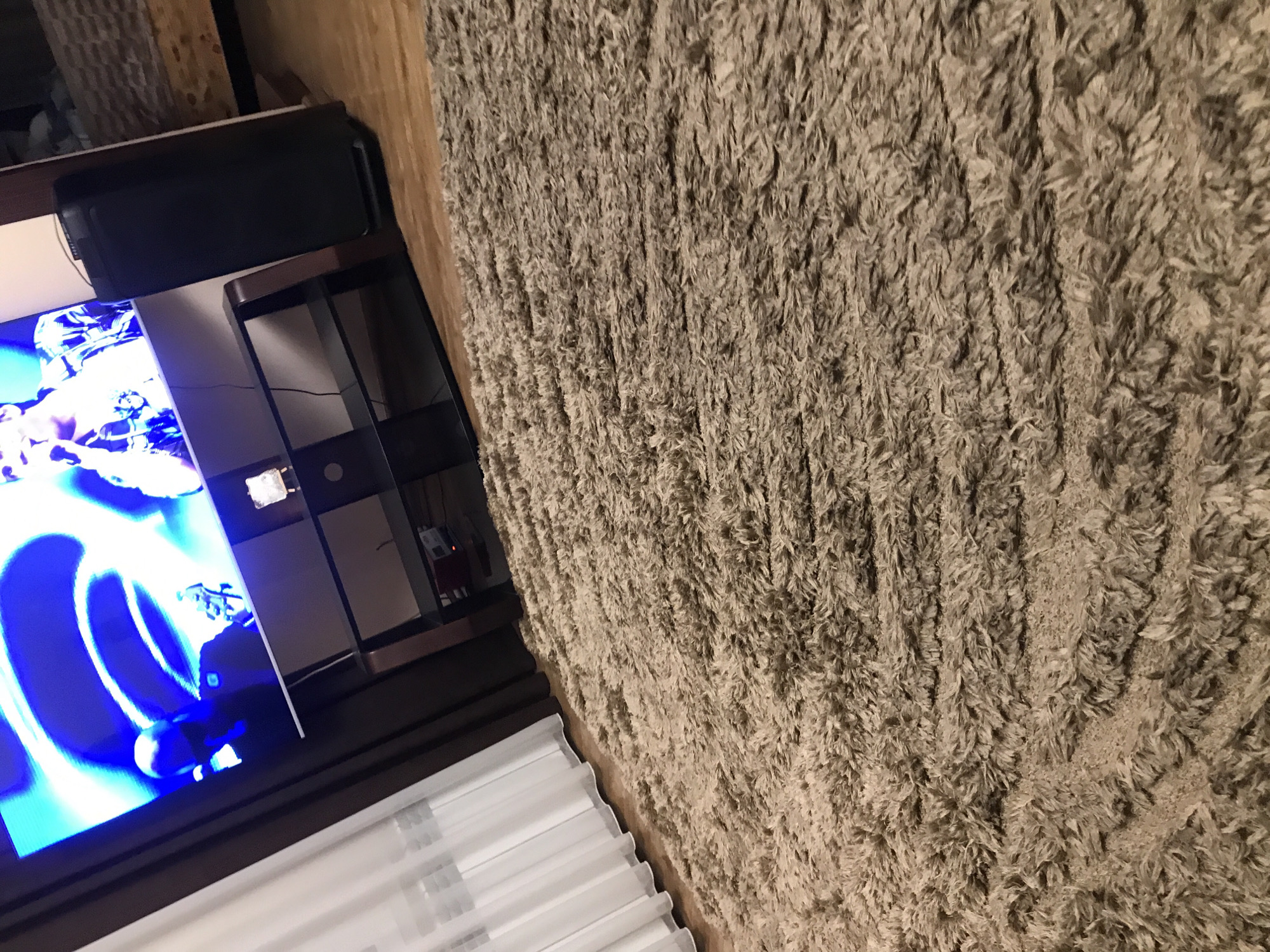 A close-up, rotated view shows a television displaying a bright blue and white image next to a multi-tiered shelving unit. A black soundbar sits atop the unit. Adjacent to the shelving unit is a light brown shag rug. The lower edge of the view includes a white window covering, possibly blinds.