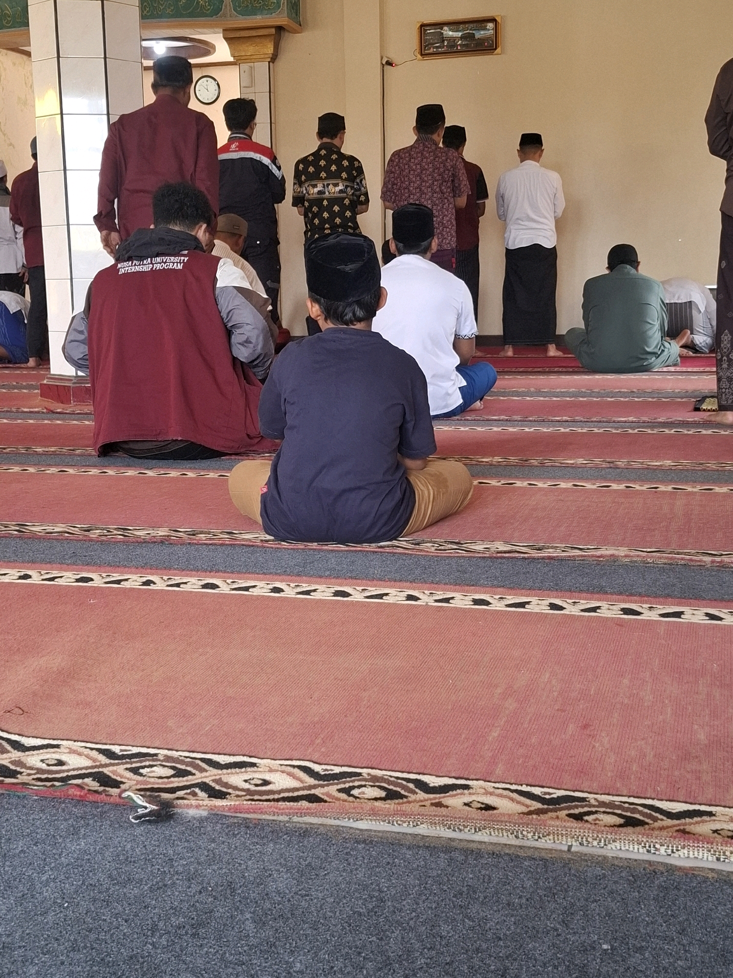 The image depicts a group of men engaged in prayer within a mosque in Ciheulang Tonggoh, Indonesia. The men are positioned on a red carpet with intricate patterned borders, facing an unseen direction of prayer. Most of the men are wearing traditional Islamic attire, including peci (caps) and sarongs, with some also wearing long-sleeved shirts or jackets. One individual in the foreground wears a maroon vest with text that appears to read "NUSA PUTRA UNIVERSITY INTERNSHIP PROGRAM." The mosque interior is visible, with a wall in the background and a clock mounted on it. The lighting suggests it is daytime. The overall scene conveys a sense of religious observance and community.