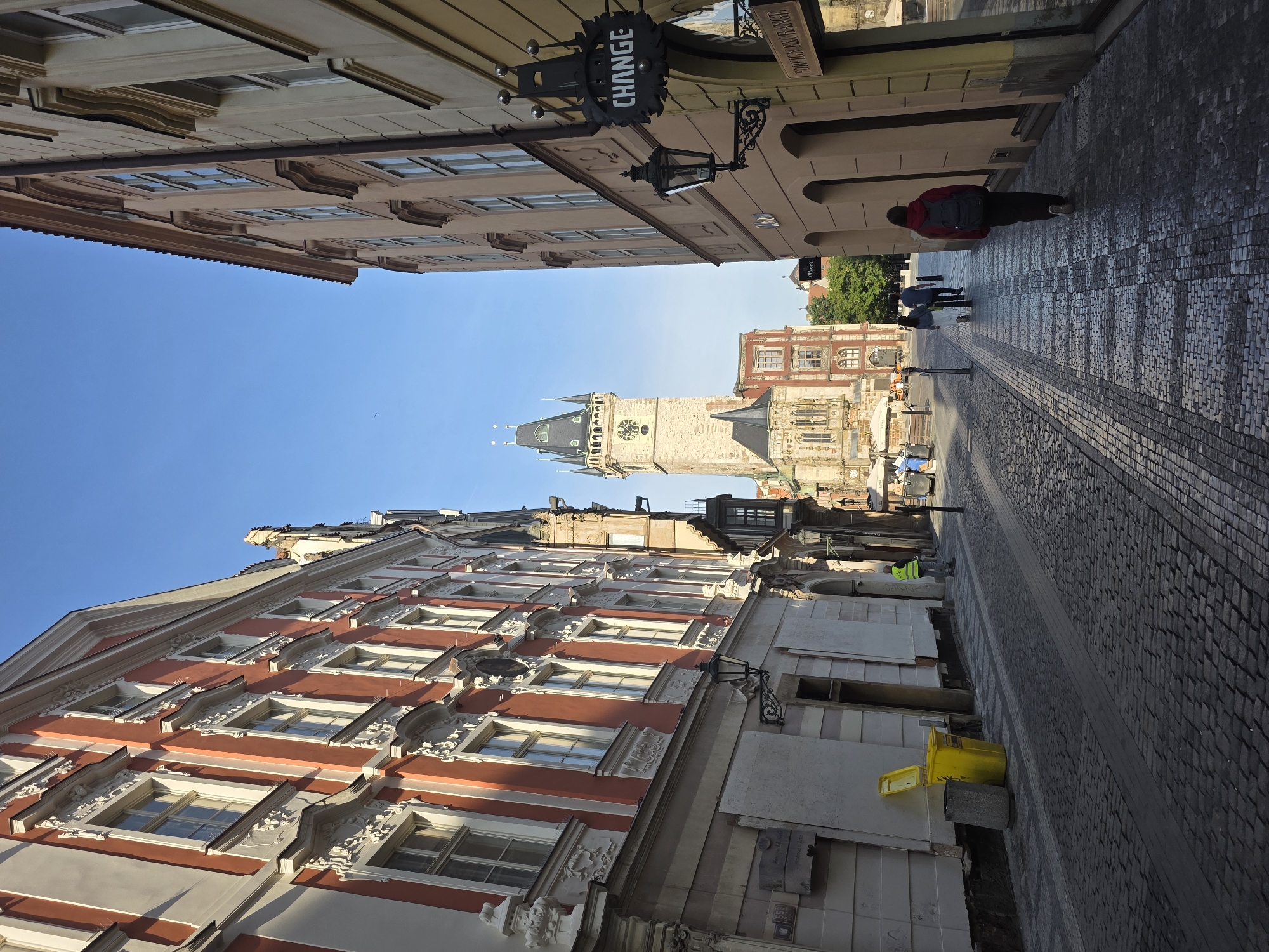 This image, viewed upright, captures a narrow, historic cobblestone street leading towards a prominent clock tower under a clear, bright blue sky. The scene is framed by multi-story buildings adorned with classical architectural details.
On the left, a richly decorated building with red and white facades features numerous windows with ornate sills and cornices. Closer to the foreground on this side, a bright yellow open bin and a grey cylindrical bin are visible at the base of the building.
On the right, the street is bordered by a lighter-colored building. A black, wrought-iron sign with white capital letters clearly spells "CHANGE" hangs from this building. A traditional black street lamp also extends from its facade. Further down the street, another faded sign with partial text, possibly "PRAGUE...", is visible.
Several people are walking away from the viewer, heading towards the distant clock tower. The closest individual on the right wears a red jacket and a grey backpack. Other pedestrians are scattered further along the street.
The focal point in the distance is a grand, light-colored clock tower topped with a distinctive dark, spired roof, indicating a significant historical landmark. It is flanked by other old buildings. The strong, clear sunlight suggests it is daytime, possibly morning or late afternoon, given the shadows cast on the cobblestone street. The overall setting is that of a picturesque European urban environment.