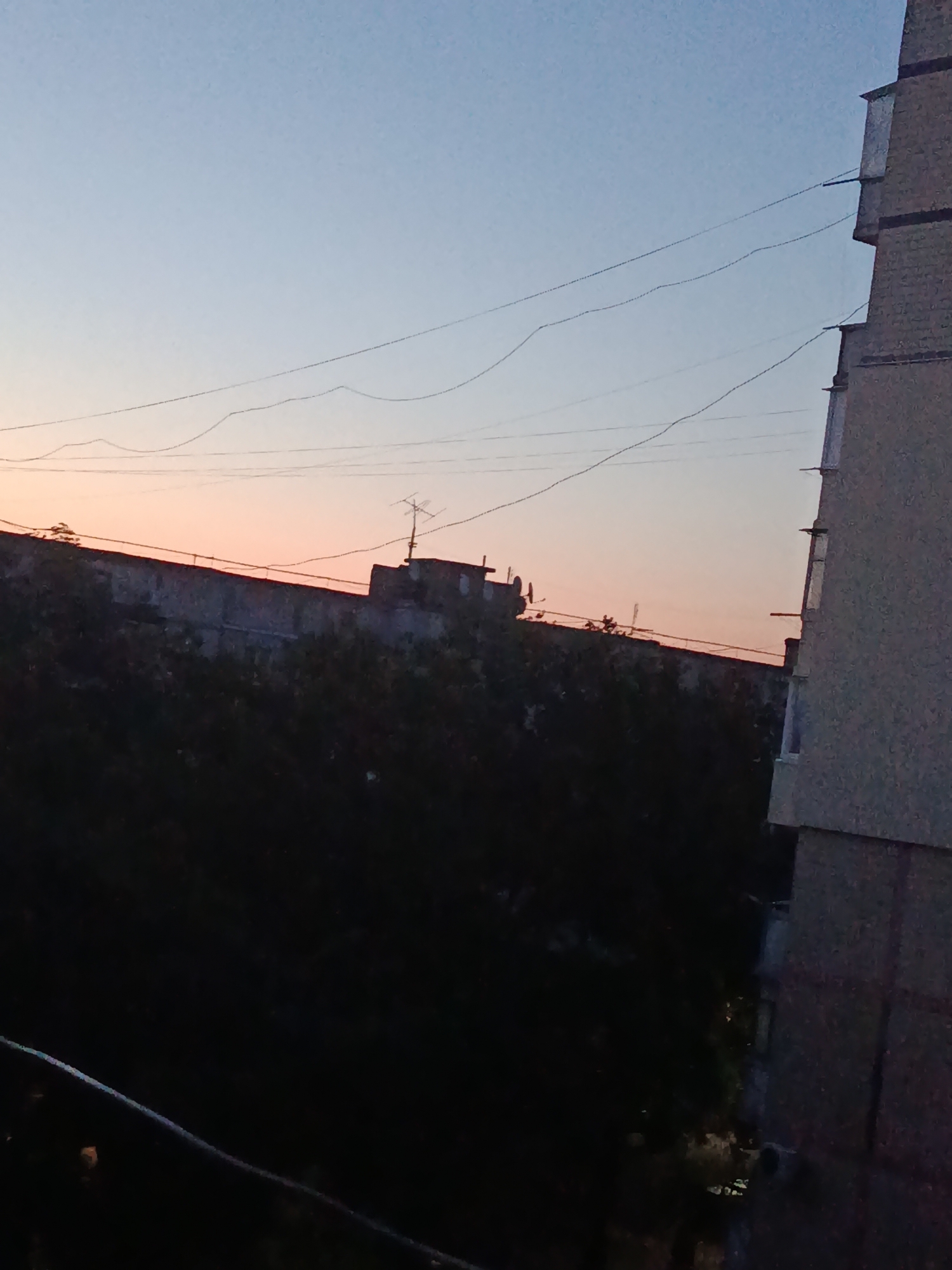 The image shows a dusk sky over Cherkaske, Ukraine. A multi-story building occupies the right side of the frame, with several power lines stretching across the sky. In the mid-ground, a long, low building with an antenna on top is visible. Trees obscure the lower part of the scene, creating a dark, silhouetted effect. The sky transitions from a pale blue at the top to a soft orange and pink near the horizon.
