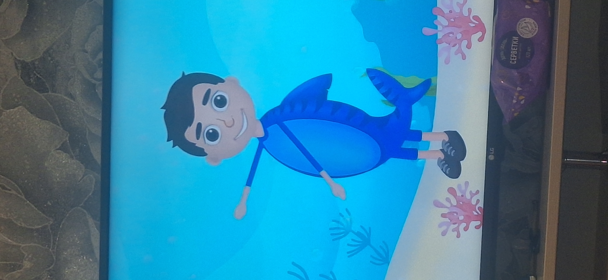A cartoon of a boy dressed as a shark is displayed on a television screen. The boy has black hair, large eyes, and a smiling expression. He is wearing a blue suit that resembles a shark costume, complete with fins and a tail. His arms are outstretched, and he is standing on a sandy seabed with coral. The background is a bright blue, representing water. Below the television screen, a shelf holds a package of wet wipes with Russian text. The image is taken indoors, and the top portion of the frame shows a textured wall. The location is not specified in the image, and therefore cannot be determined.