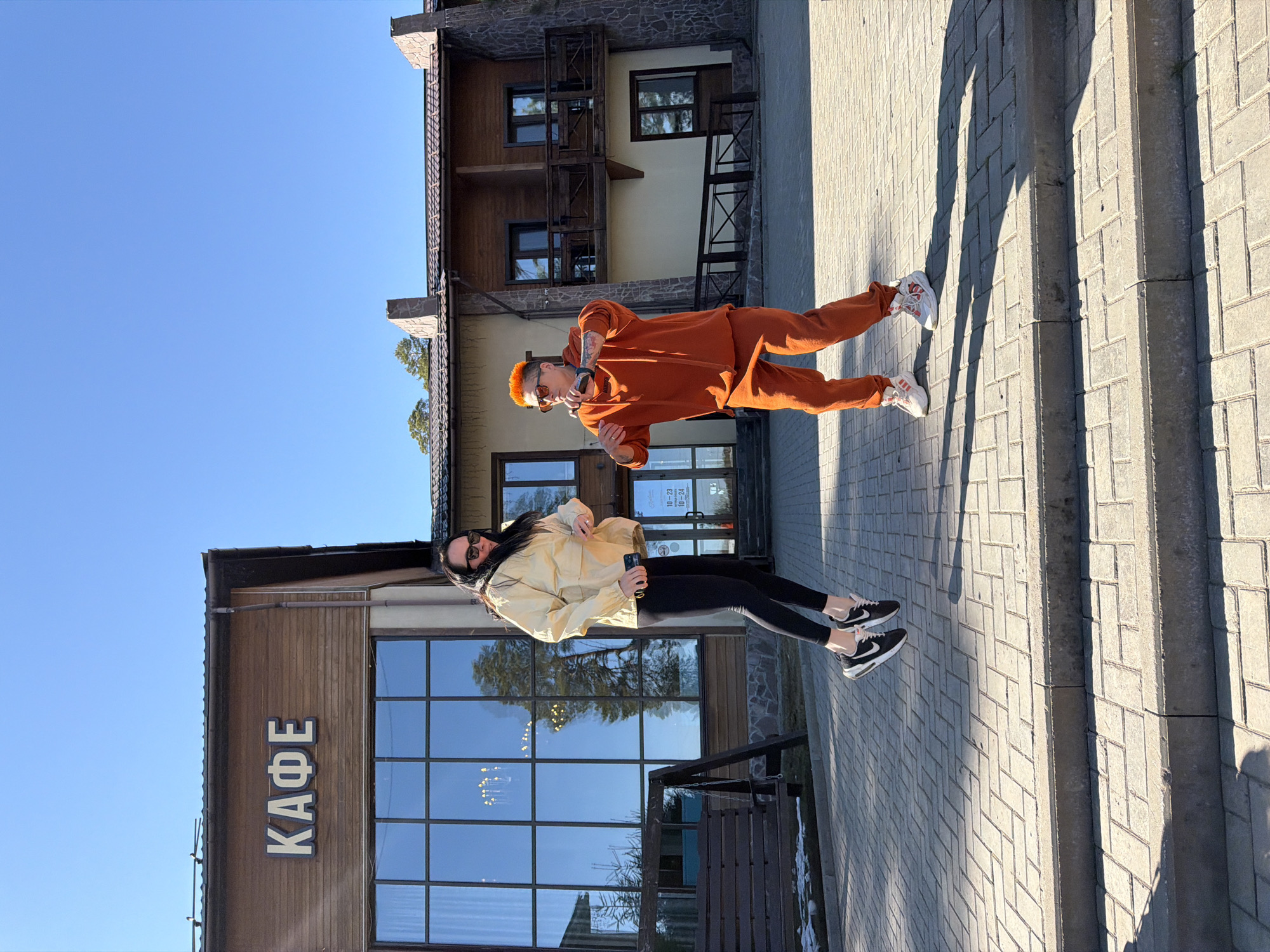 Two individuals are on a paved area in Votkinsk, Russia. One individual is wearing an orange sweatsuit and orange tinted glasses, posing with their fingers to their temple. The other individual is wearing a light yellow jacket and black leggings, facing the opposite direction. Behind them is a building labeled "КАФЕ," indicating a cafe. Further in the background is another building with multiple windows and balconies. The sky is clear and blue.