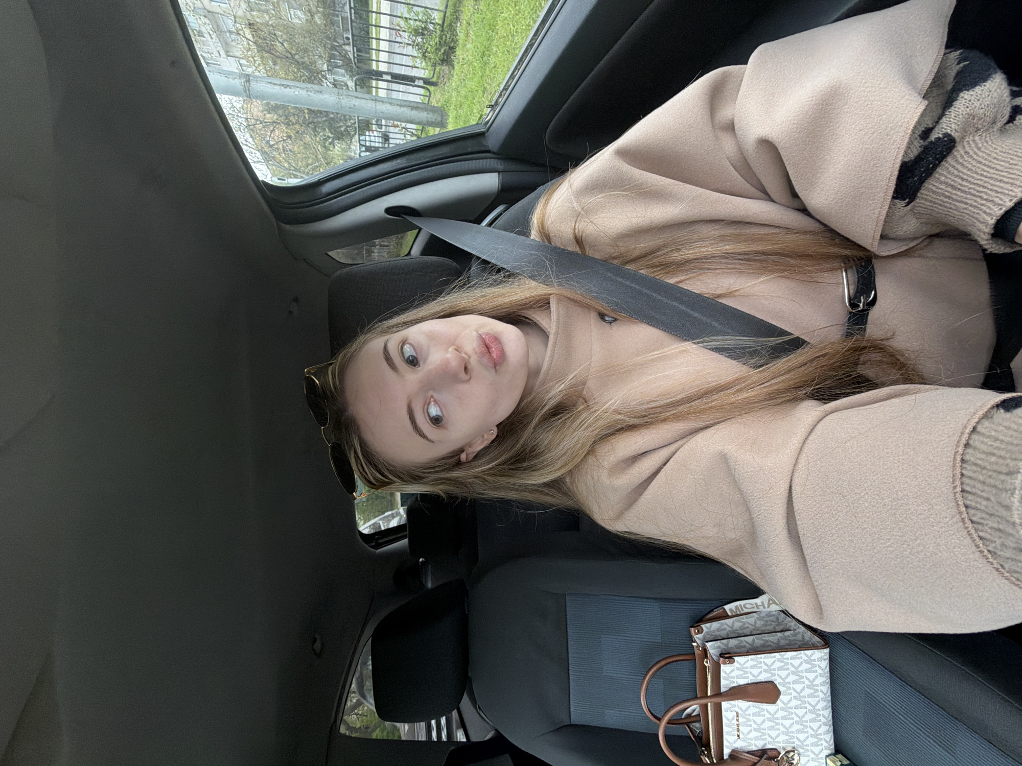 A young woman is seated in the driver's seat of a car, wearing a beige coat and a seatbelt. She is looking at the camera with a neutral expression. Her blonde hair is long and partially covers the seatbelt. A purse with a white and brown design is on the passenger seat. The background shows a street scene with trees and a building, hinting at an urban environment. The location appears to be in Plovdiv, Bulgaria, as suggested by the prompt. The image captures a casual moment inside a vehicle.