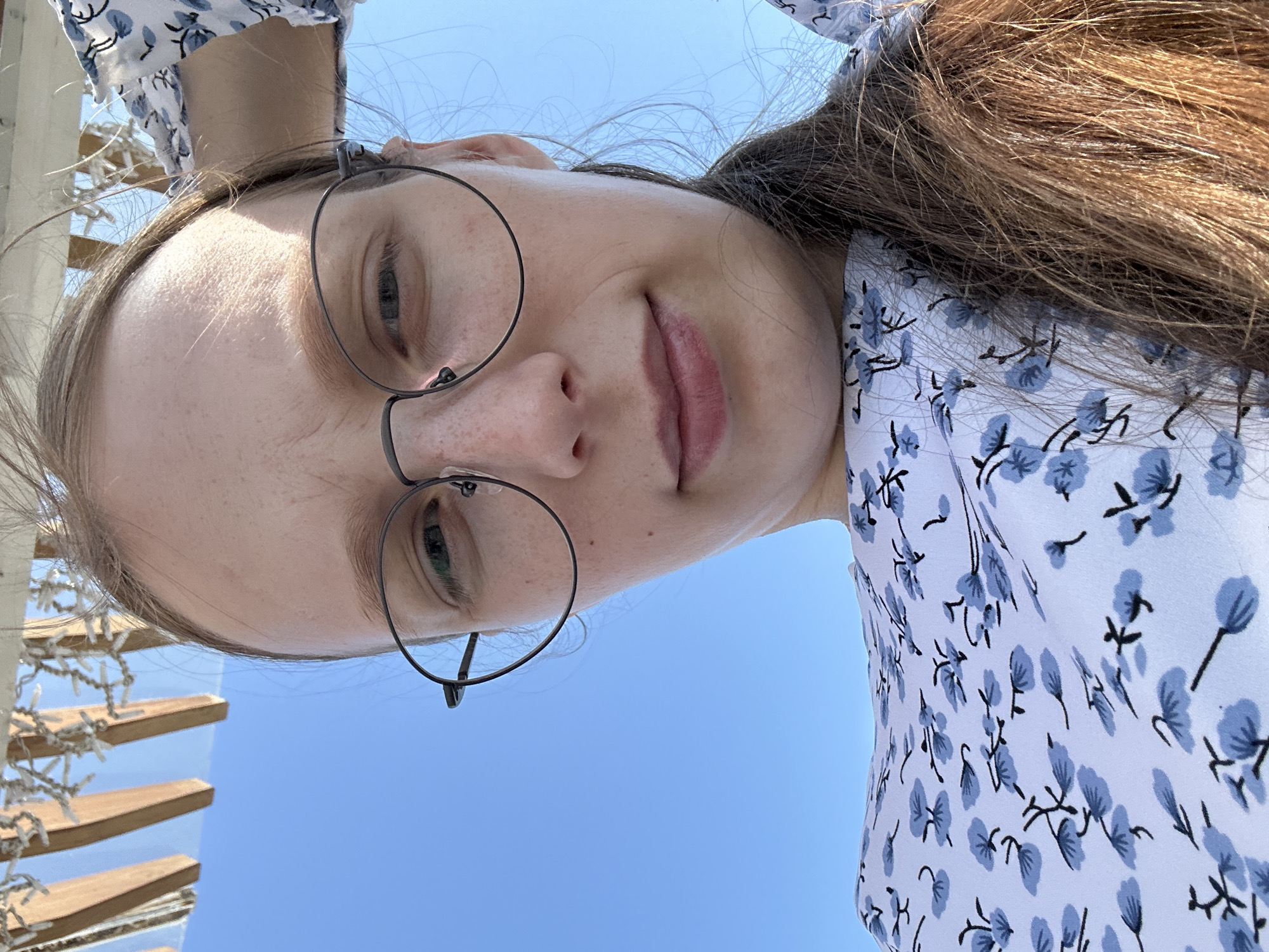A close-up, low-angle selfie of a woman with long brown hair, wearing round glasses and a white dress with a blue floral pattern. She is outdoors with a clear blue sky in the background and what appears to be a wooden structure with lights overhead. The woman's right hand is resting on the back of her head. The setting is a sunny day, possibly in Металлплощадка, Russia.