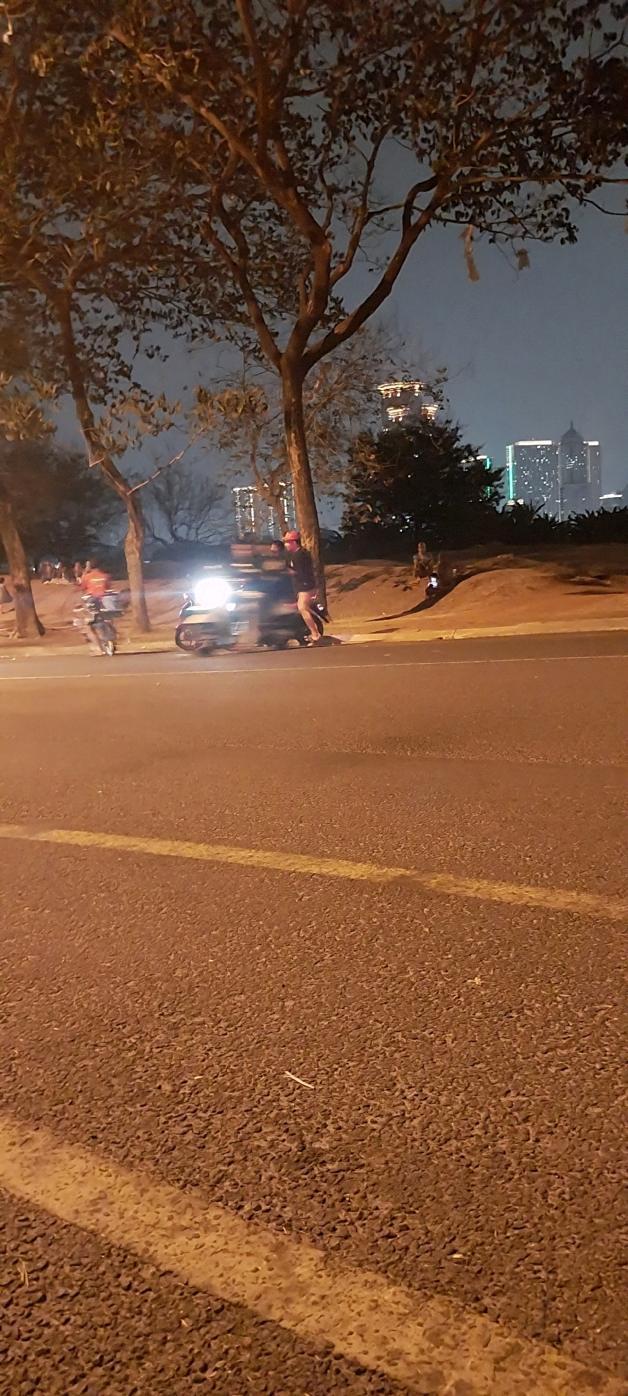 The image depicts an outdoor scene at night, likely in a city like Kelapa Dua, Indonesia, given the urban skyline in the background. A paved road occupies the foreground, marked with a yellow line. To the left, several trees are visible, their branches sparsely leafed.
In the middle ground, a person wearing a red hat and dark clothing stands next to a scooter. The scooter's headlight is illuminated, casting a bright light. Further to the left, another person is visible on a motorcycle, also with a headlight on. In the background, a cluster of modern buildings with illuminated windows forms a city skyline against the dark night sky. The terrain slopes upwards behind the road, covered with sparse vegetation and a few trees. The overall atmosphere suggests a public space at night, possibly a roadside park or promenade.