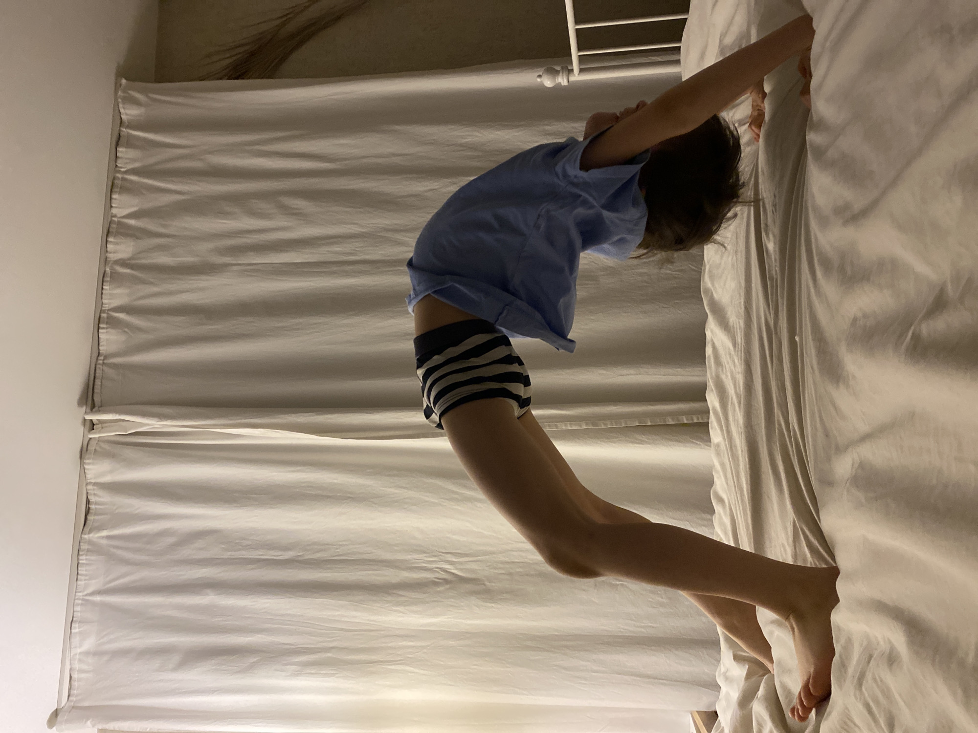 A child is performing a backbend on a white bed. The child is wearing a light blue t-shirt and striped shorts. The bed has white sheets and is next to a white wall. A white curtain hangs to the left of the bed. A portion of a white metal bed frame is visible at the top of the image. The location is not identifiable as Yekaterinburg, Russia.