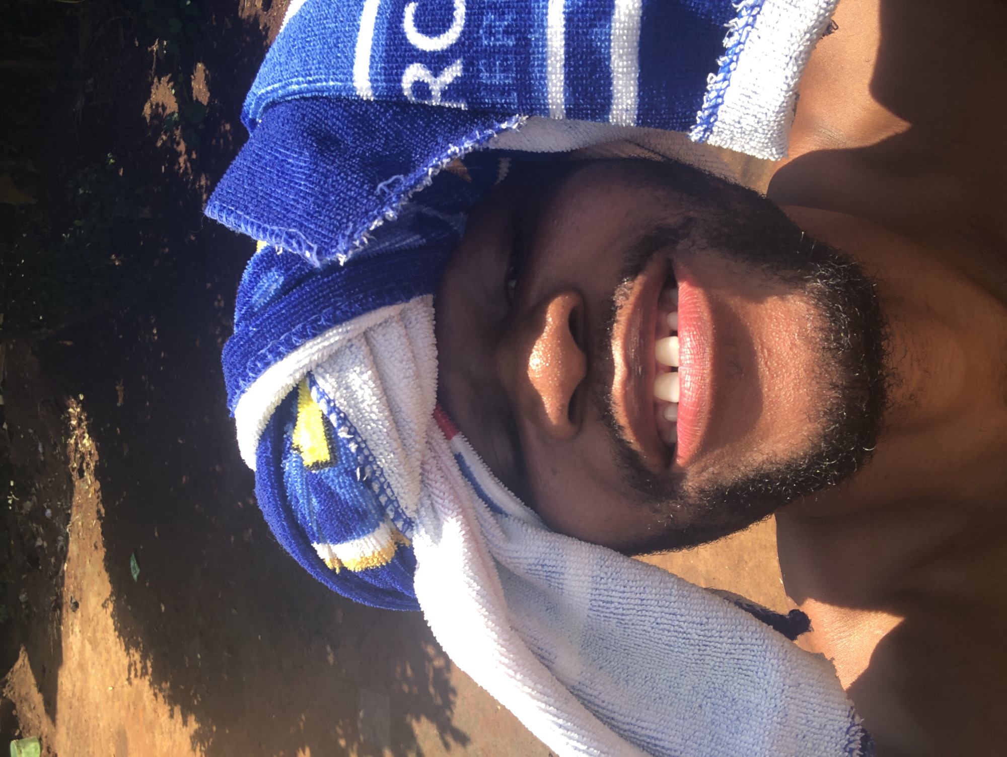 A man smiles while wearing a blue and white striped towel on his head. The location is Unknown, Unknown.