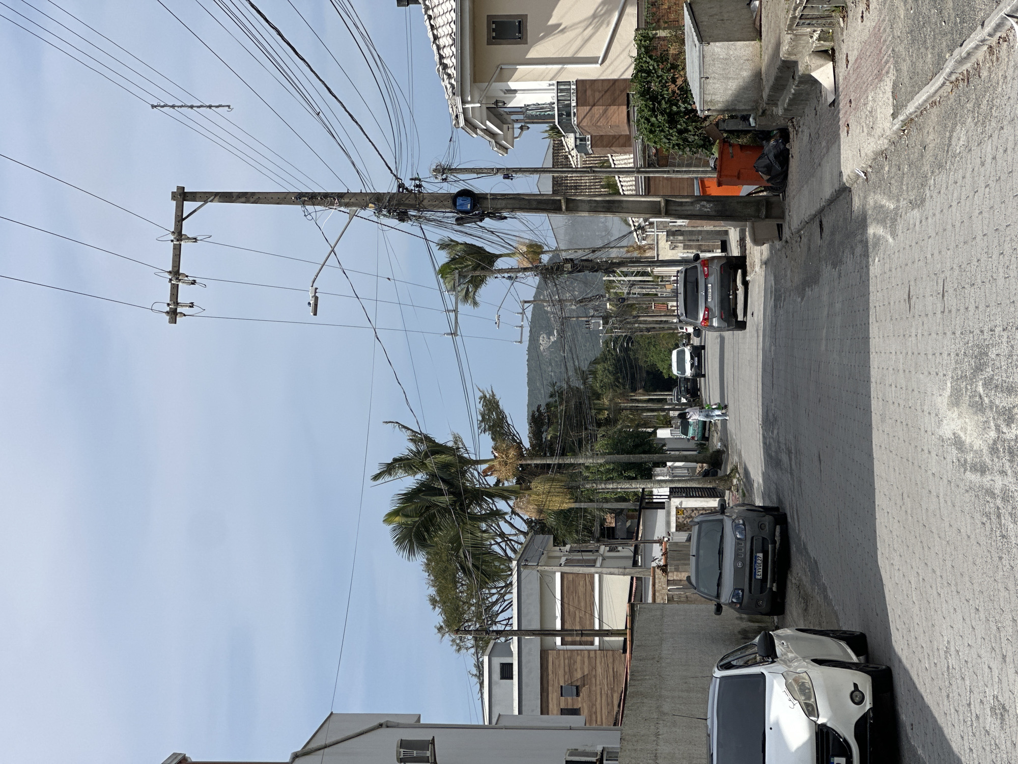 The image depicts a street scene in Florianópolis, Brazil. A paved road, constructed with grey cobblestones, runs horizontally through the frame, lined with residential buildings and utility poles. Several vehicles are parked along the street and in motion. On the left, a white hatchback is parked near the foreground, followed by a grey Fiat Uno. Further down the road, a grey SUV is visible. A person in white clothing walks on the sidewalk on the right side of the street, pulling a cart. Utility poles with numerous wires are a prominent feature, crisscrossing the sky. One pole has a blue device mounted on it. Residential buildings, with a mix of modern and older architectural styles, flank both sides of the street, some featuring balconies and tiled roofs. Vegetation, including palm trees and other greenery, is present throughout the scene. In the background, a hilly landscape with scattered trees is visible under a hazy sky. An orange trash bin and a pile of black trash bags are situated on the right side of the road.