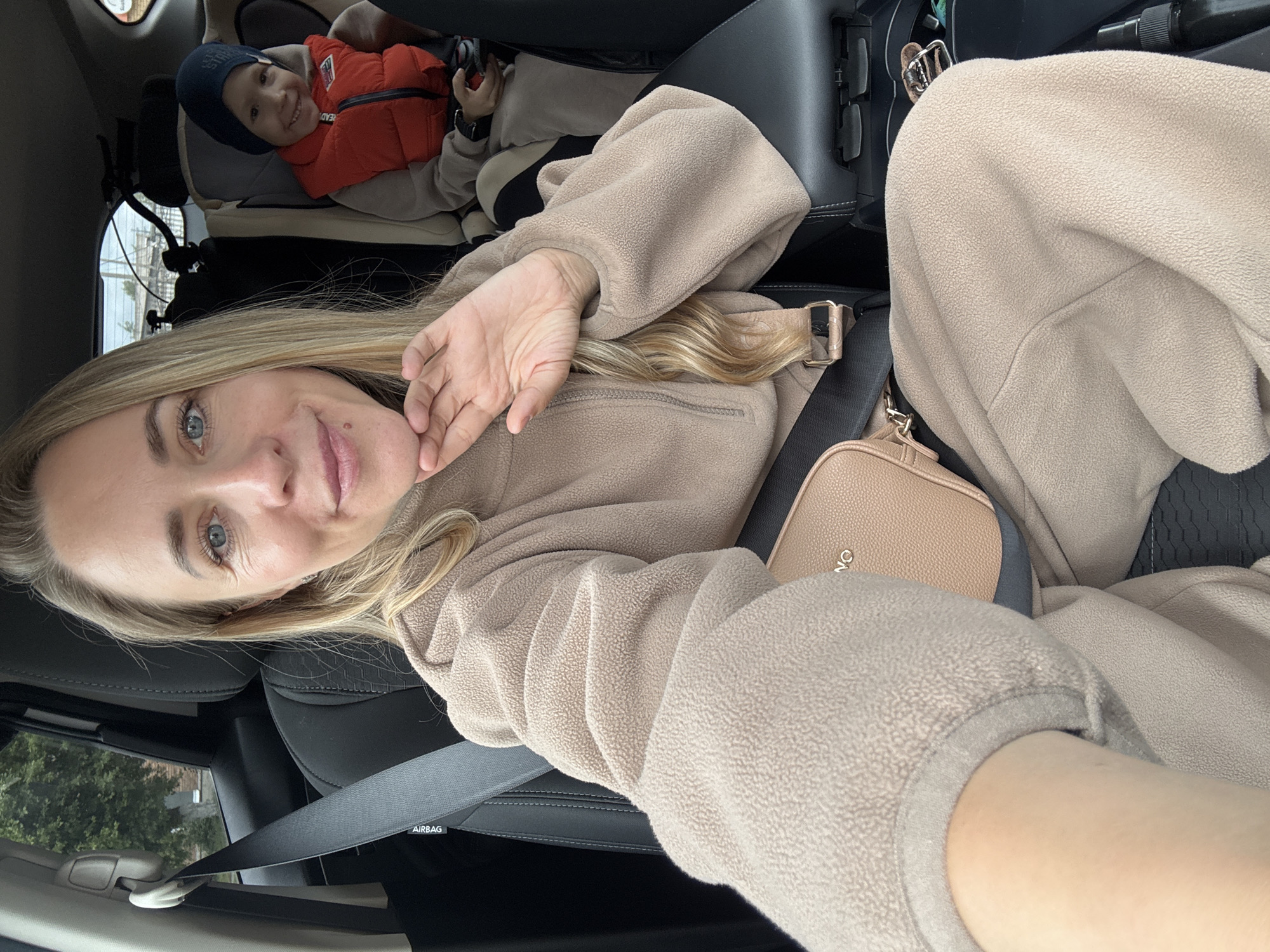 A woman is taking a selfie in a car. She is wearing a beige fleece jacket and has a beige purse across her body. She is wearing a seatbelt. A child in an orange puffer vest and blue hat is in a car seat in the background. The location may be Chaikovsky, Russia.
