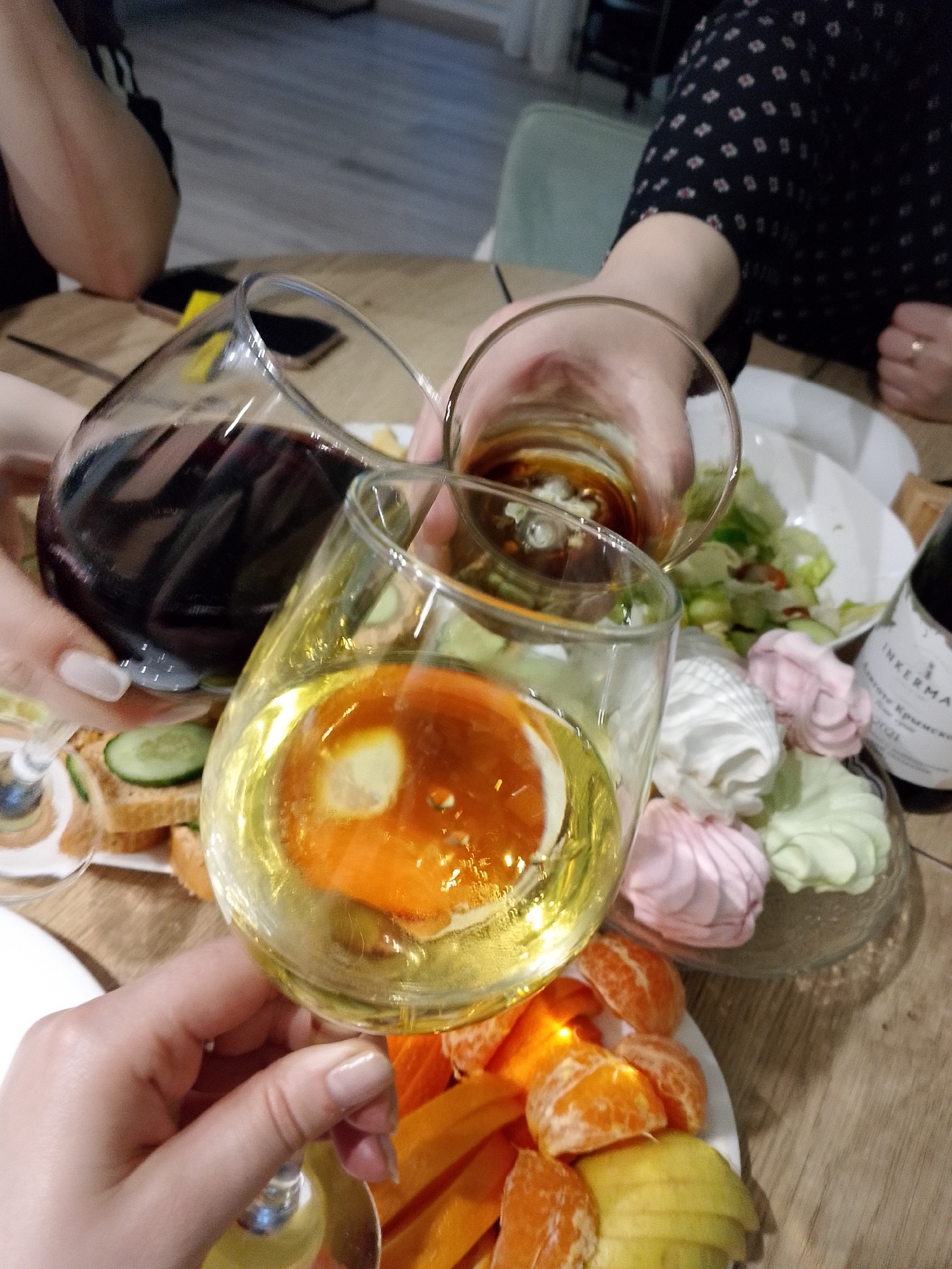 The image shows a close-up view of a celebratory scene on a wooden table. Three hands holding glasses are prominent: one contains red wine, another contains a dark amber liquid, and the third contains white wine with a slice of orange.
The table is set with plates of food, including sandwiches with cucumber, a salad, marshmallows (pink, white, and green), and sliced fruit (tangerines, persimmons, and pears). A bottle of "INKERMAN" Crimean wine is also visible on the table.
The setting appears to be indoors, possibly in a home or restaurant. No specific information confirms the location as Izhevsk, Russia, beyond the linguistic origin of the wine bottle.