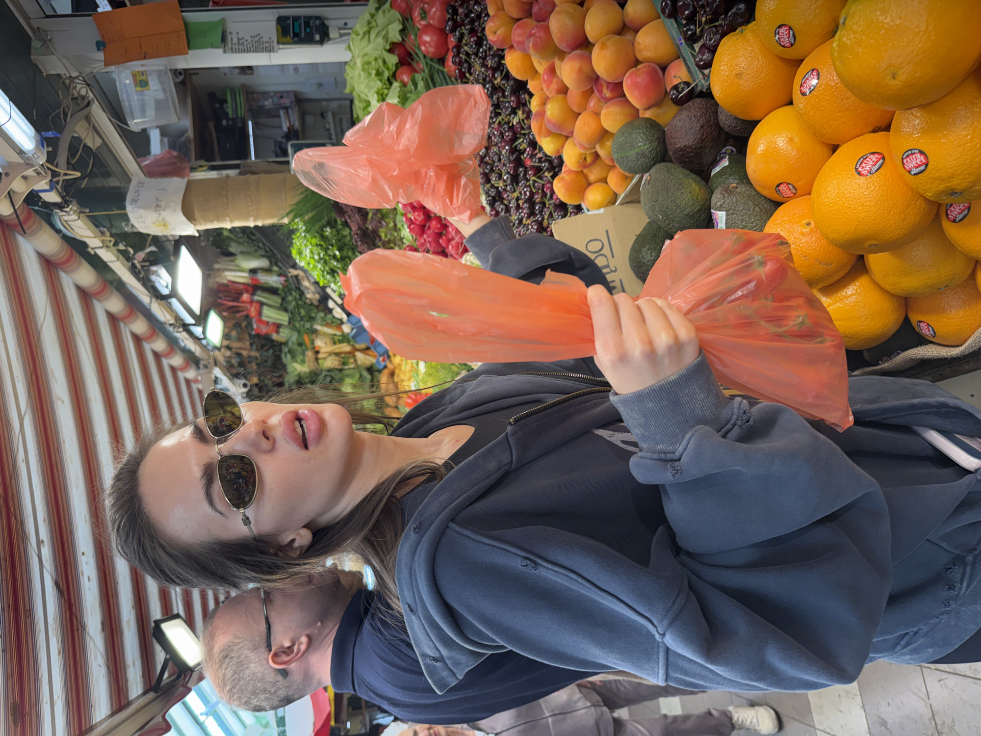 A medium, candid shot shows a woman and a man standing in what appears to be a fresh produce market. The woman, who is in the foreground, is wearing sunglasses and a gray hoodie. She is holding two orange plastic bags, likely filled with groceries. The man, partially visible behind her, is wearing glasses and a dark blue shirt.
The background is filled with various fruits and vegetables. Oranges, avocados, and cherries are stacked on the right side of the frame. To the left, there are green leafy vegetables and red tomatoes. The market appears to be well-lit with fluorescent lights.
The presence of an awning suggests an outdoor market setting. The location is not specified, but the presence of market stalls and fresh produce indicate an urban environment.