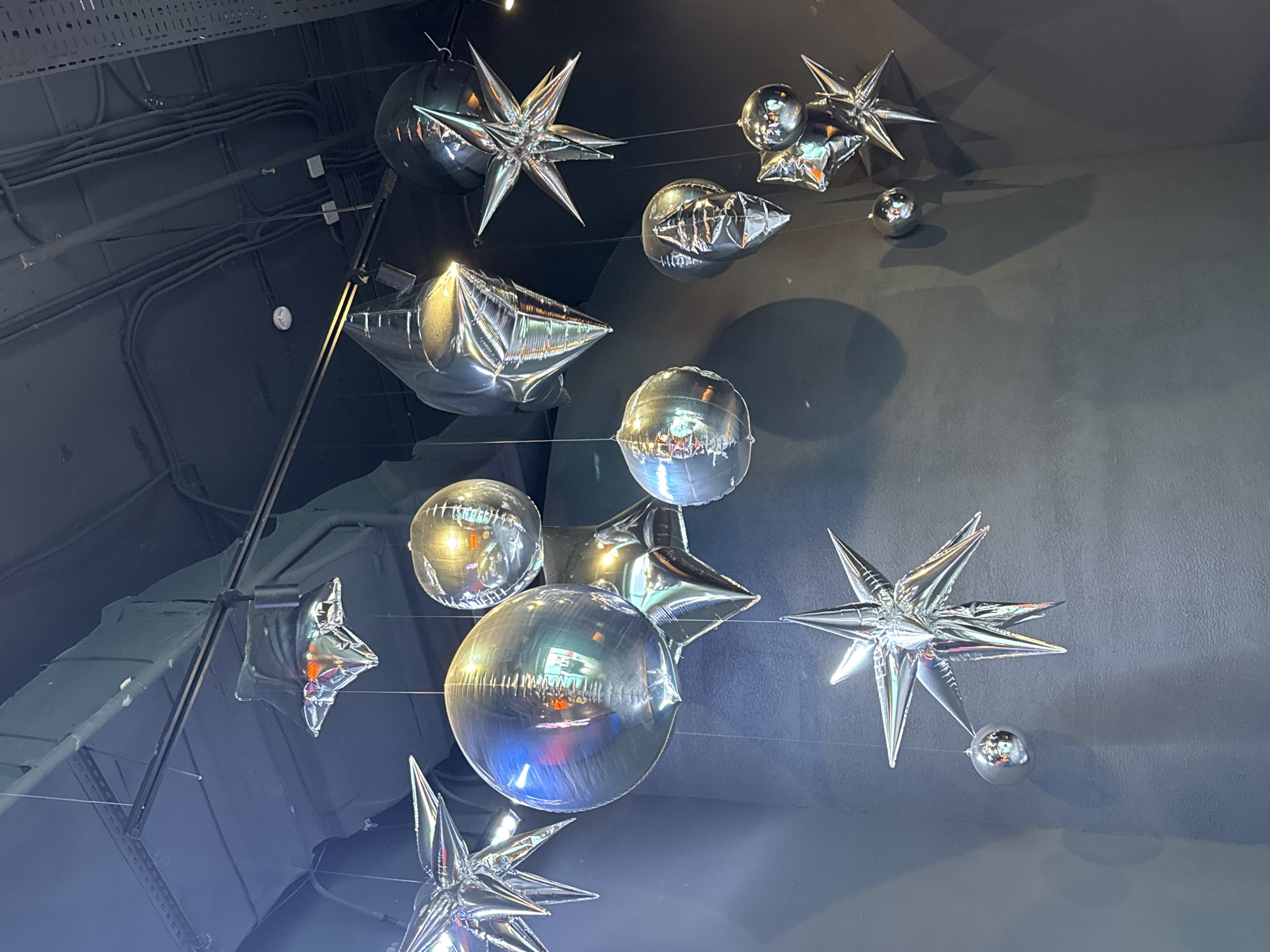 The image depicts a collection of silver foil balloons hanging from the ceiling. The balloons are of varying shapes, including spheres and stars. They appear to be suspended by thin wires or strings. The background is a dark, possibly black or gray, ceiling with exposed pipes and other fixtures. The balloons reflect light, creating bright highlights. The image was taken indoors, and no people are visible. There is no evidence to suggest the location is Saint Petersburg, Russia.