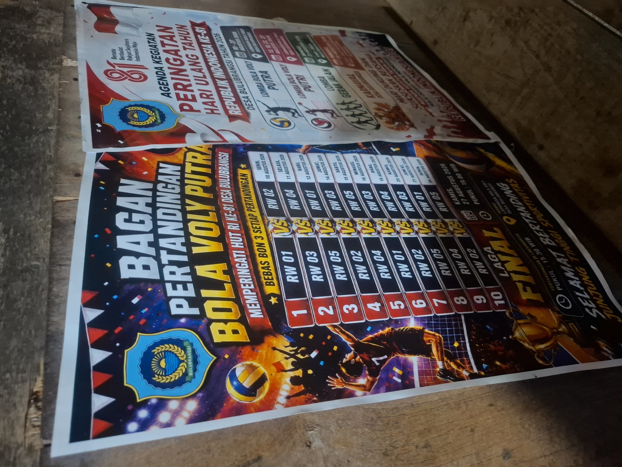 The image shows two posters displayed on a wooden surface, likely a wall or fence, in an outdoor or semi-outdoor environment with a rough wooden background.
The poster on the left is predominantly black with vibrant graphics, featuring a volleyball tournament titled "BAGAN PERTANDINGAN BOLA VOLY PUTRA MEMPERINGATI HUT RI KE-81 DESA BULUBRANGSI". It includes a schedule with match pairings and dates. A graphic of a male volleyball player in action, a volleyball, a trophy, and silhouettes of cheering spectators are visible. The crest of Bulubrangsi is also present.
The poster on the right is primarily white and red, with the title "AGENDA KEGIATAN PERINGATAN HARI ULANG TAHUN REPUBLIK INDONESIA KE-81 DESA BULUBRANGSI TAHUN 2026". It lists various events, including "LOMBA BOLA VOLY PUTRA", "LOMBA BOLA VOLY PUTRI", "LOMBA GERAK JALAN", and "KARNAVAL". The crest of Bulubrangsi is also on this poster.
The wooden surface appears weathered and rustic. The lighting suggests it is daytime. The overall scene depicts a community event poster in Bulubrangsi, Indonesia.