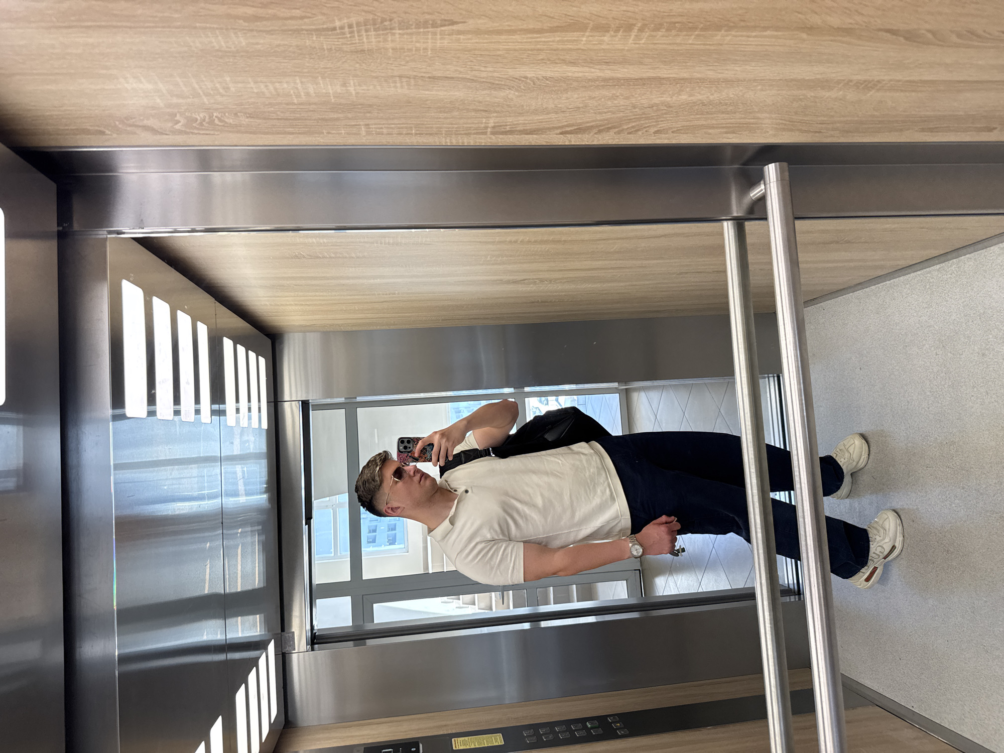 A fair-skinned man is taking a selfie in an elevator. He's wearing sunglasses, a white short-sleeve polo shirt, dark blue pants, and white sneakers. He is holding a smartphone in his left hand and wearing a watch on his left wrist. A black shoulder bag is slung across his body.
The elevator has stainless steel walls, a wood-paneled ceiling, and a grey floor. There is a stainless steel handrail and a control panel with buttons near the floor. A window behind the man reveals a view of a building with multiple windows, suggesting an urban setting.
There are no distinctive architectural features to identify the specific location within Plovdiv, Bulgaria.