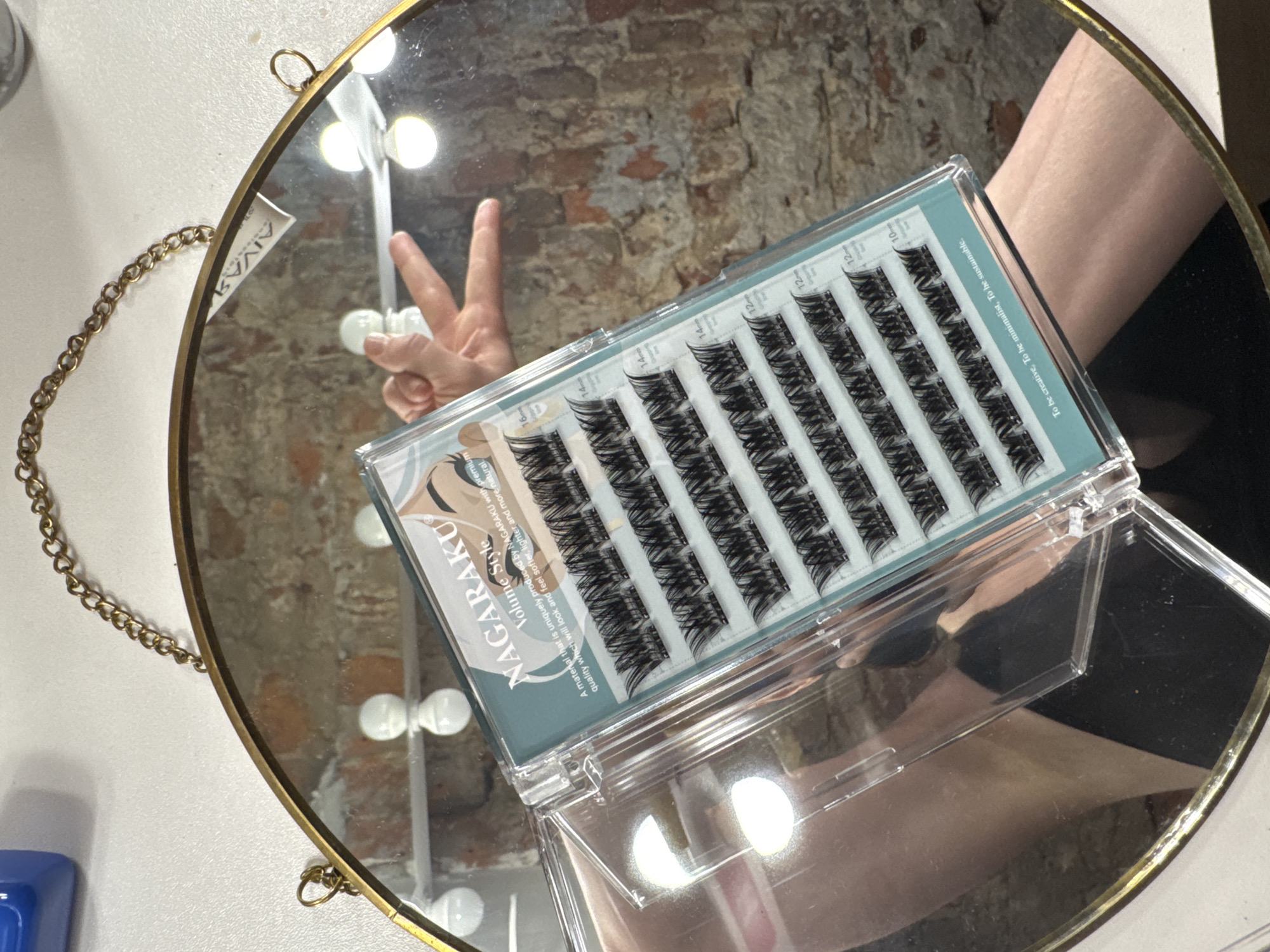 The image is an overhead shot of a vanity mirror with a gold frame and chain. The mirror reflects a hand giving a peace sign. An open package of NAGARAKU Volume Style individual false eyelashes is on the right side of the mirror. The package contains multiple rows of lashes. A vanity light is reflected in the mirror behind the hand. The background is a brick wall. The location of the image is Saint Petersburg, Russia.