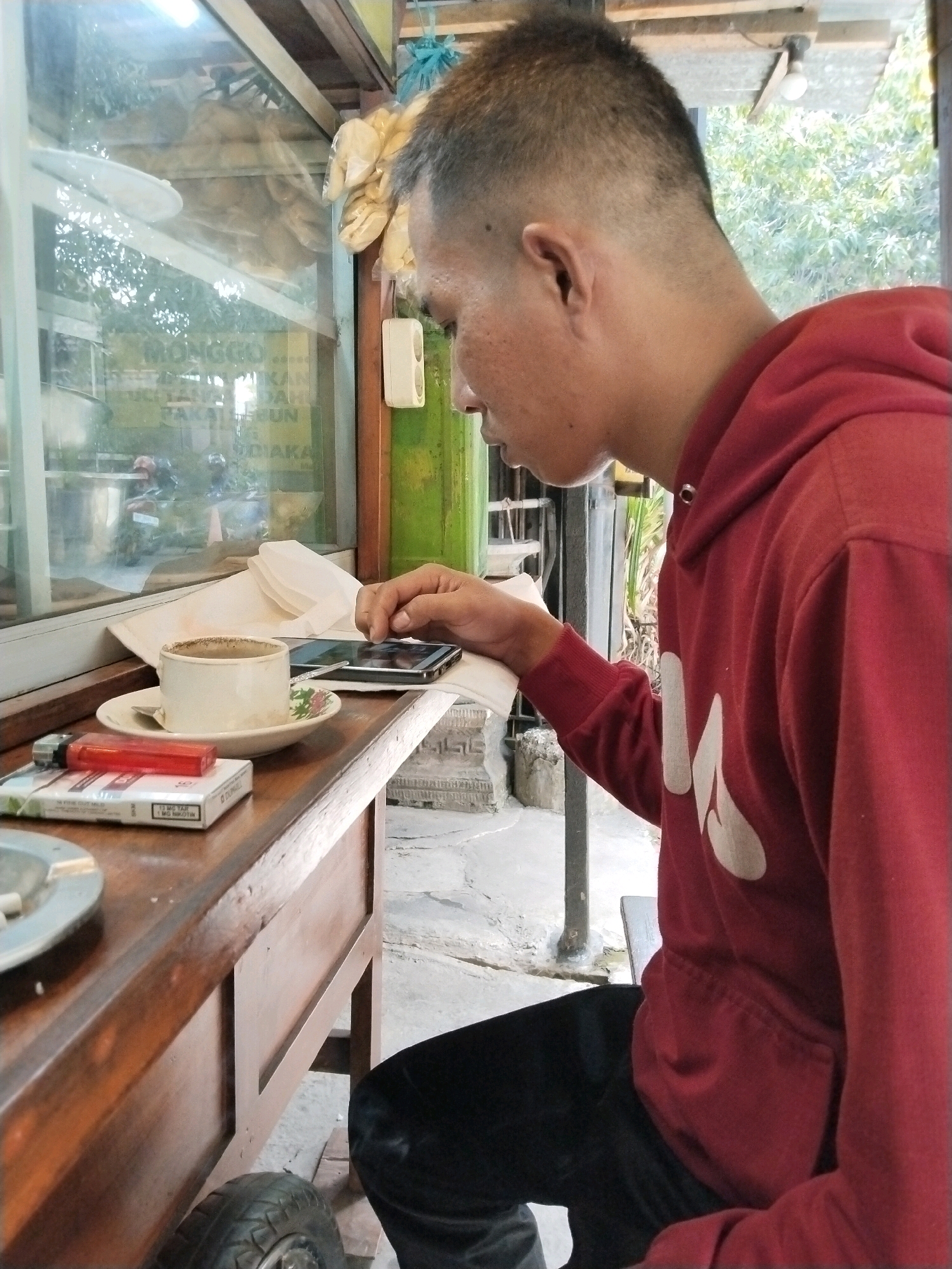 A man in a red hooded sweatshirt and black pants is sitting at a wooden table, looking down at his phone. On the table in front of him are a cup of coffee on a saucer, a cigarette pack, and a lighter. The scene appears to be outdoors, with a window behind the man showing reflections of trees and a sign in Indonesian that reads "MONGGO... SILAHKAN MAKAN PAKAI SABUN..." which translates to "PLEASE... PLEASE EAT USING SOAP..." This phrase seems nonsensical, possibly a humorous or ironic sign. The man is also interacting with his phone, his finger touching the screen. The environment is a bit rustic, with a visible wheel attached to the table's base and an ashtray on the table. The overall impression is of a casual moment at a roadside eatery or cafe. The location is identified as Trenggalek, Indonesia, which aligns with the Indonesian language on the sign.